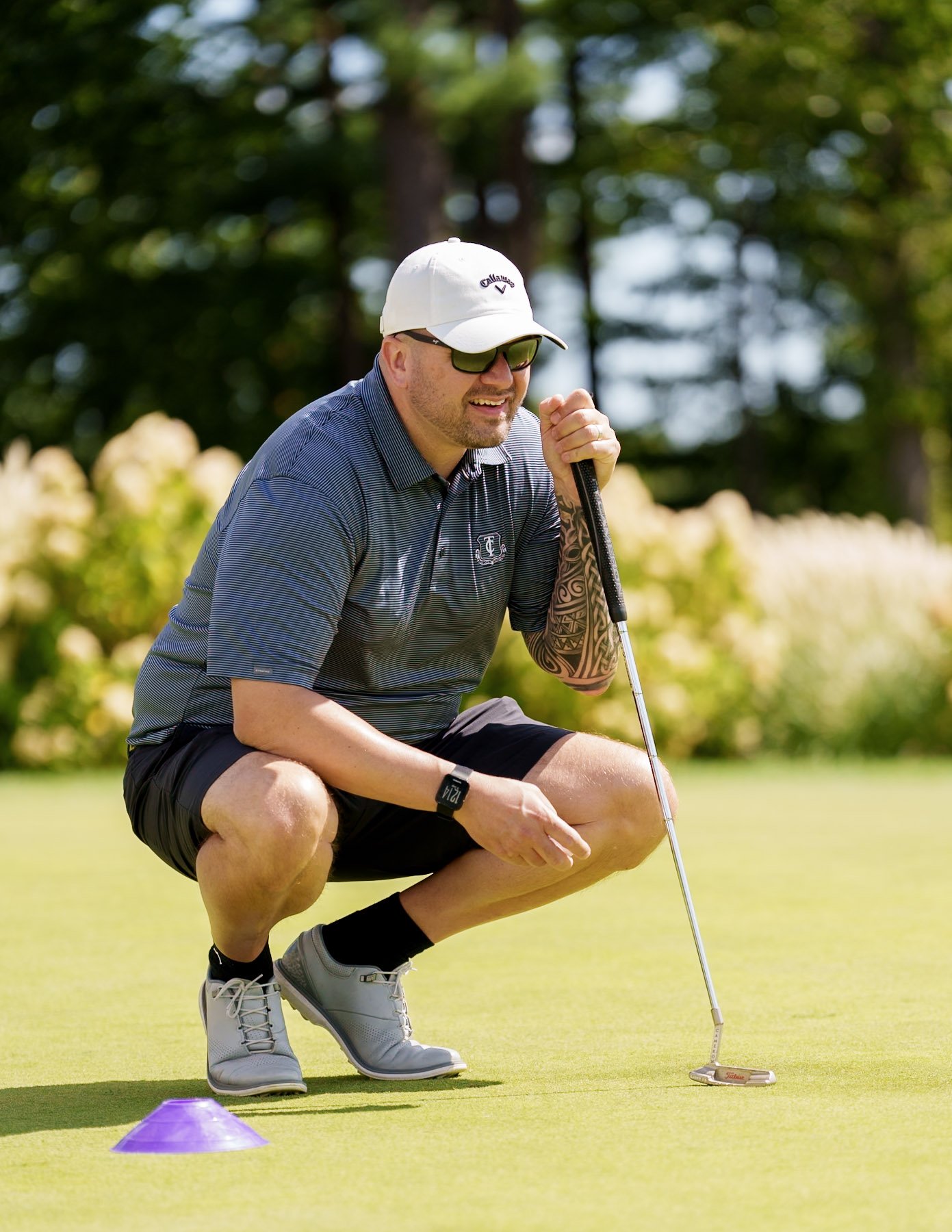 A man crouching on a golf course, holding a golf club, preparing to make a putt. He's wearing a white cap, sunglasses, a gray striped polo shirt, black shorts, and gray golf shoes. There is a purple golf tee marker on the green grass.