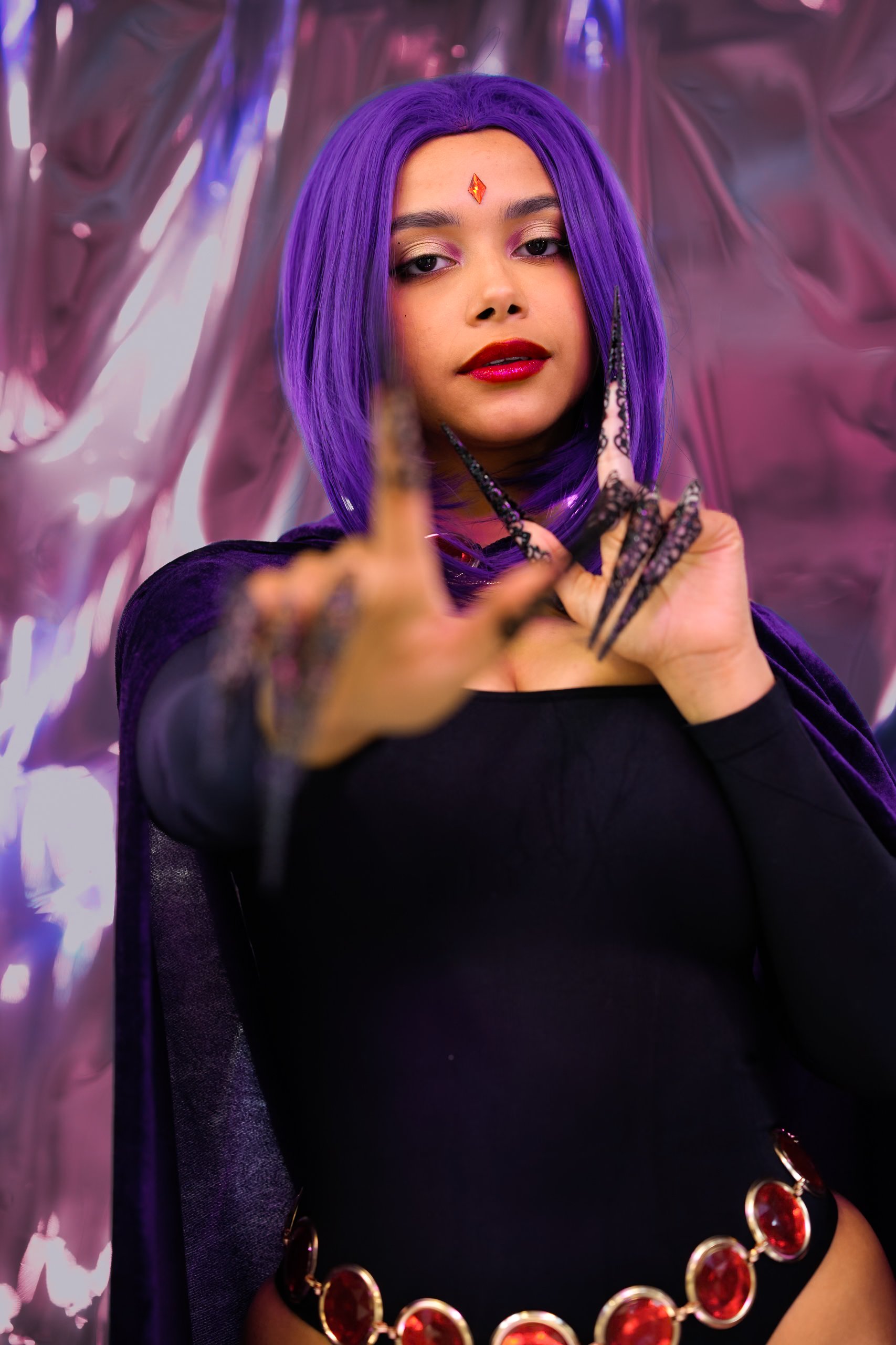 Young woman with purple hair, wearing black outfit with a jeweled belt, purple cape, and black lace claws, posing with her hand extended forward in front of a pink and purple metallic backdrop.