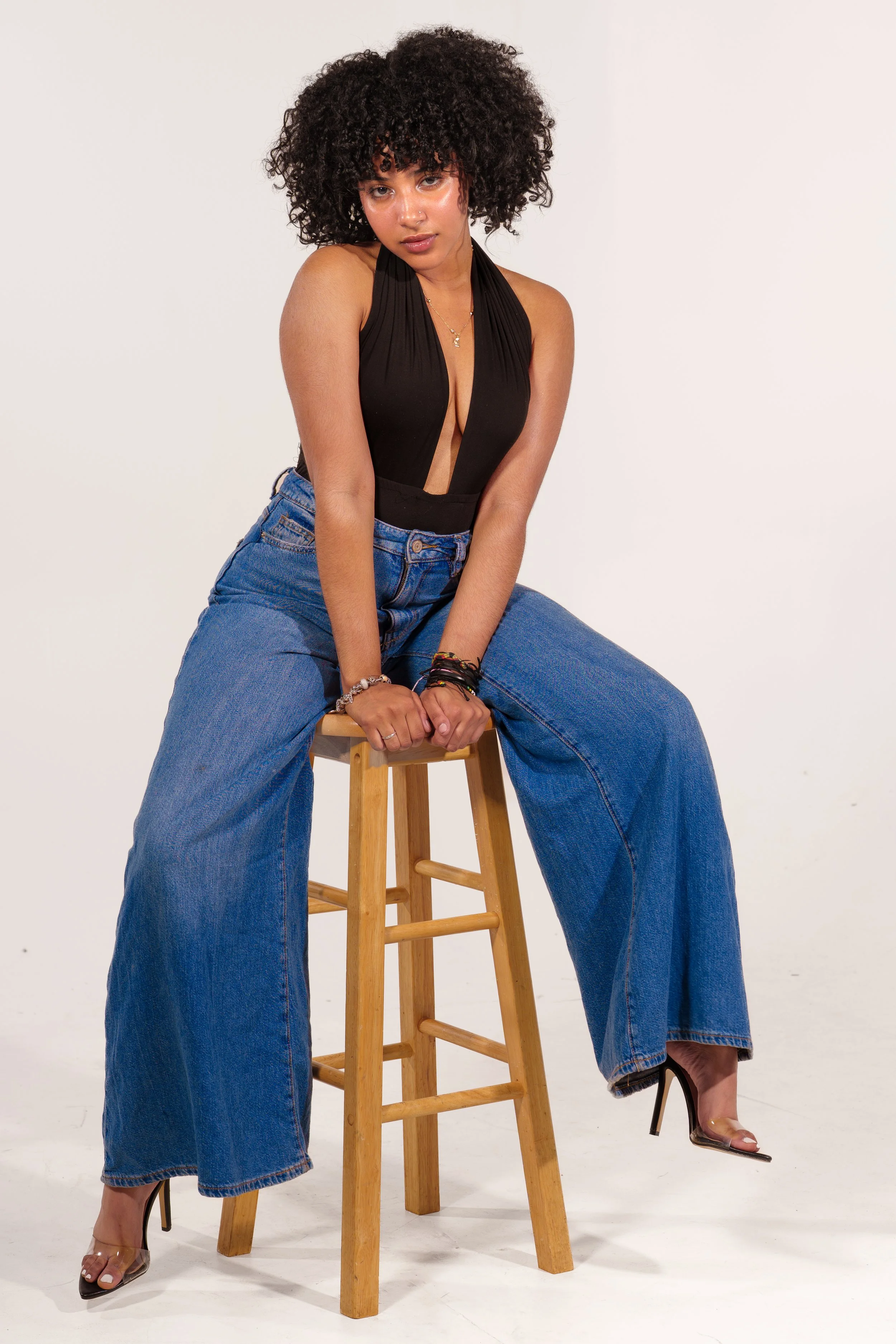 A woman with curly hair sitting on a wooden stool against a plain white background, wearing a black sleeveless top, wide-leg jeans, and high heels.
