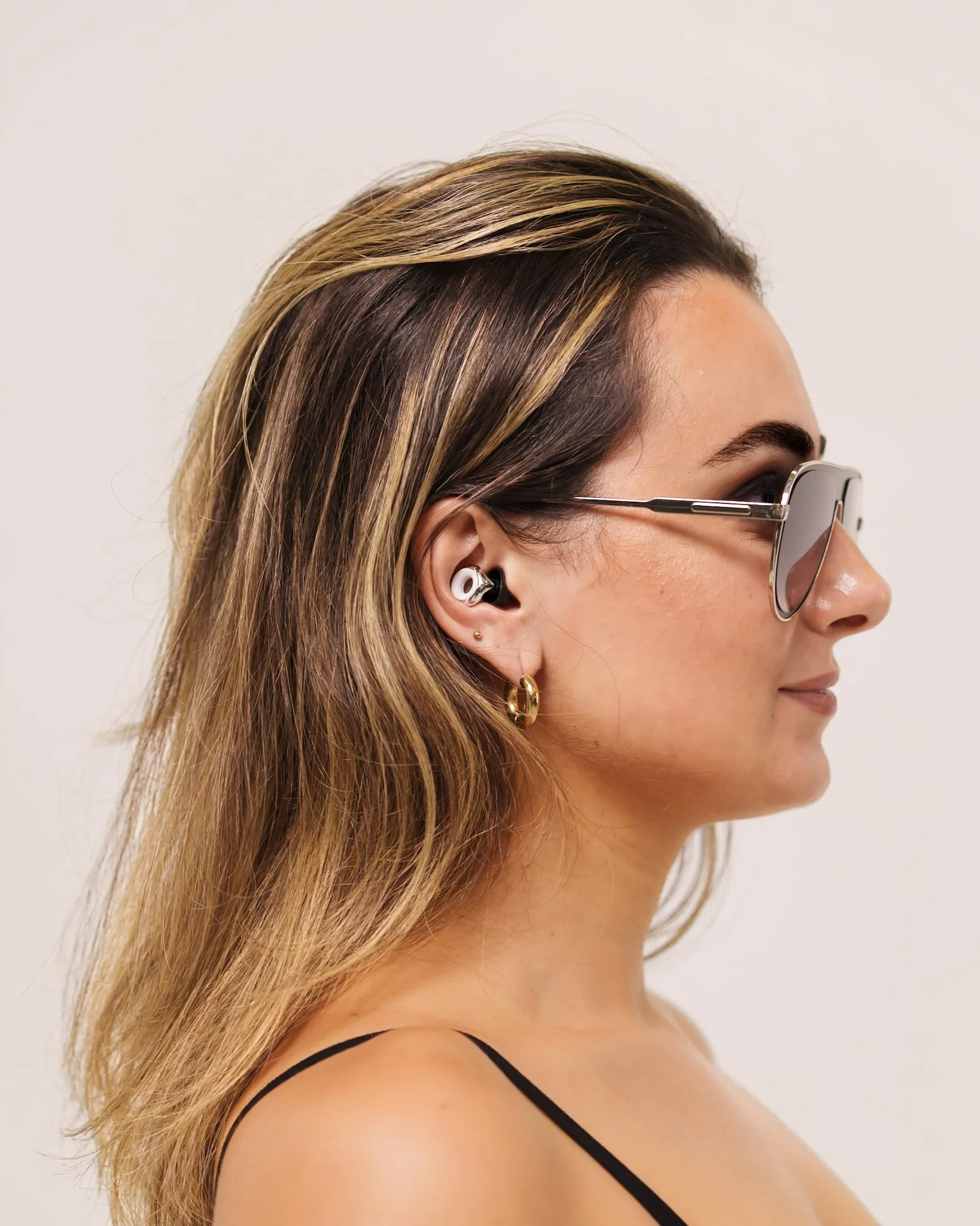 A woman with light brown hair and highlights wearing sunglasses, gold hoop earrings, and wireless earbuds, facing right.