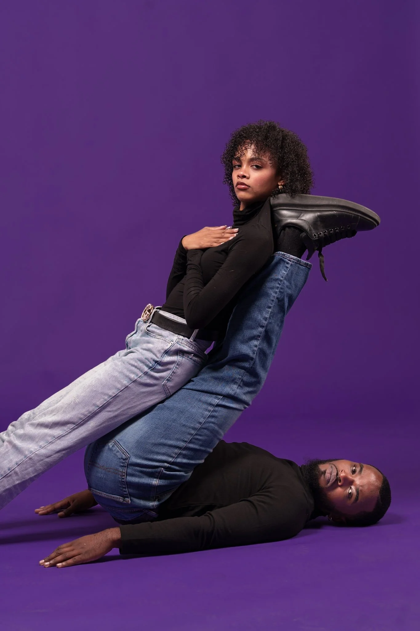 A man is lying on the floor with his legs raised, supporting a woman sitting on his shins against a purple background. The woman has curly hair, wears a black turtleneck, light-colored jeans, and black sneakers, and looks directly at the camera. The 