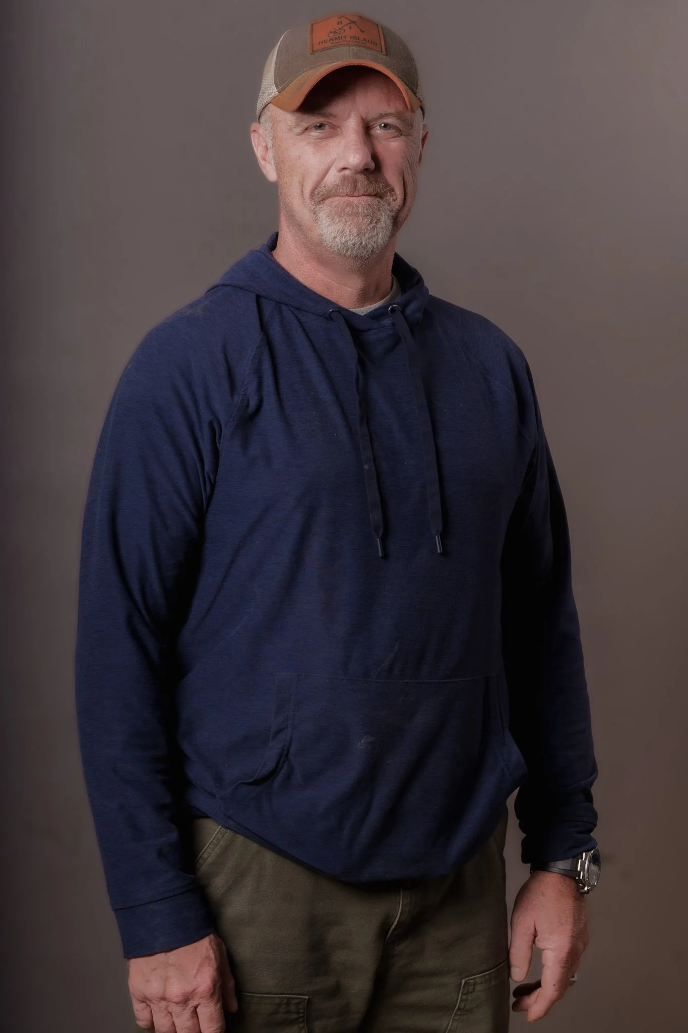 A middle-aged man wearing a beige and orange baseball cap, a navy blue hoodie, and olive green cargo pants, standing against a plain background.