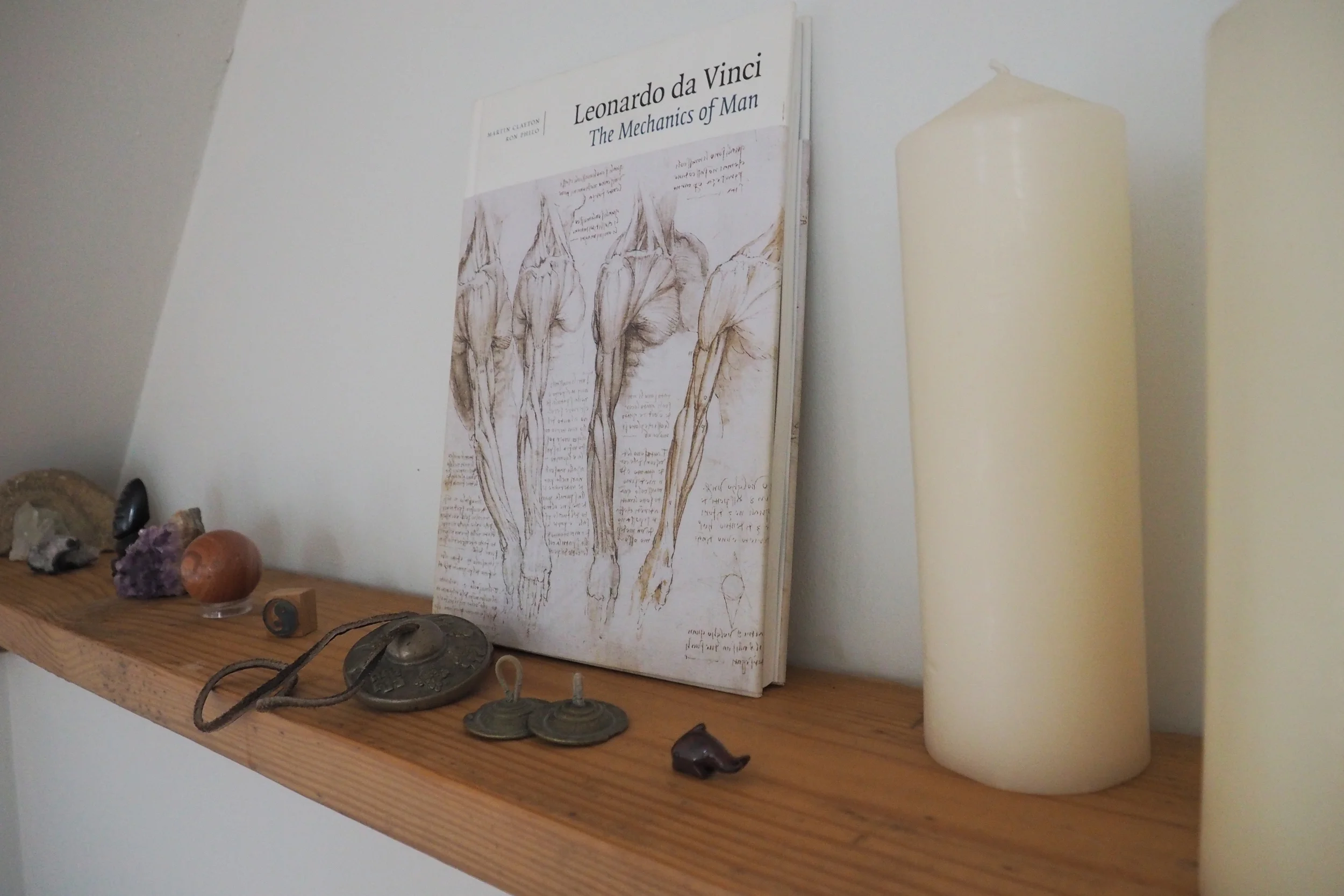 A wooden shelf with assorted objects, a book titled 'Leonardo da Vinci: The Mechanics of Man' featuring sketches of human anatomy, and two large cream-colored candles.