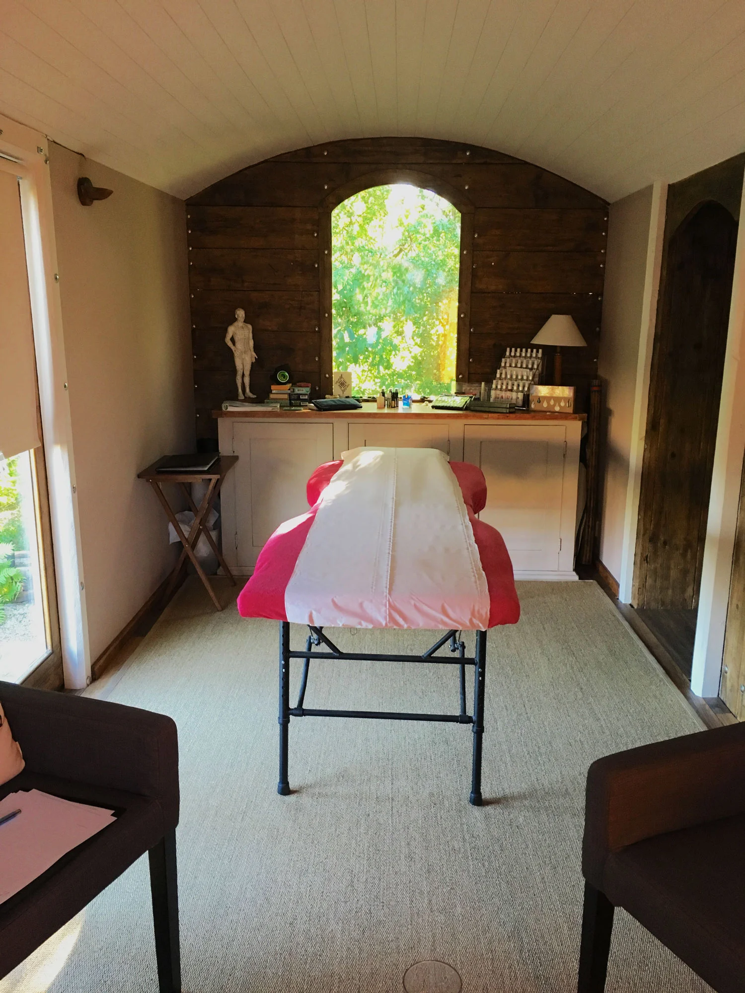 Massage table covered with a pink and white sheet in a massage room with a window revealing green foliage outside. The room has beige carpet, white and wooden walls, and a wooden ceiling.