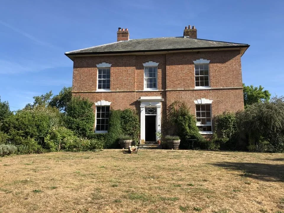 A large two-story brick house with six windows, a white front door, and a small front porch, surrounded by bushes and a lawn, under a clear blue sky.