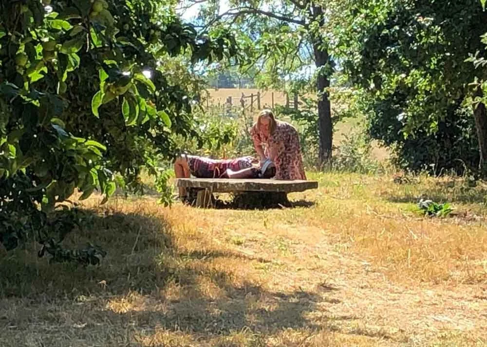 Woman lying on a wooden platform outdoors, receiving a massage from another woman in a floral dress, surrounded by trees and sunny weather.