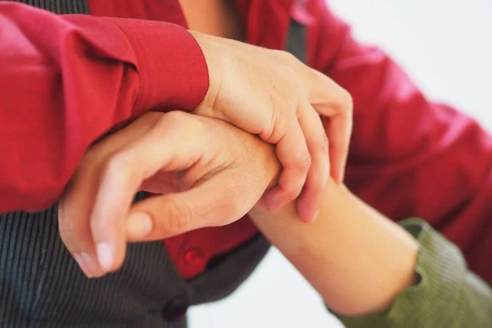 Close-up of two people holding hands, one wearing a red long-sleeve shirt and the other a green long-sleeve shirt.