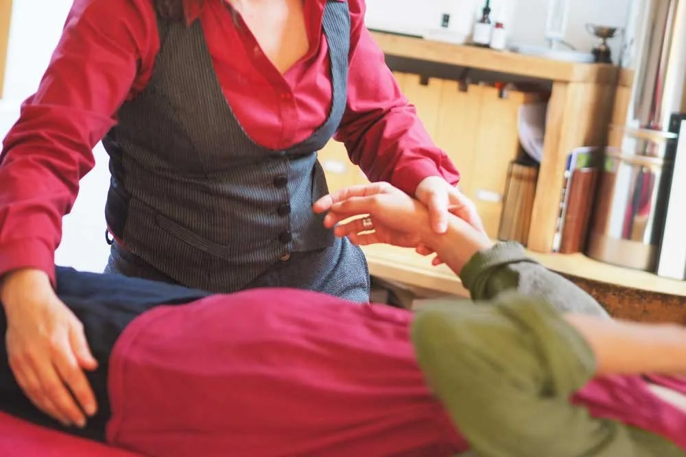 A person in a red shirt and gray vest helping someone by holding their hand in a wellness or medical practice setting.