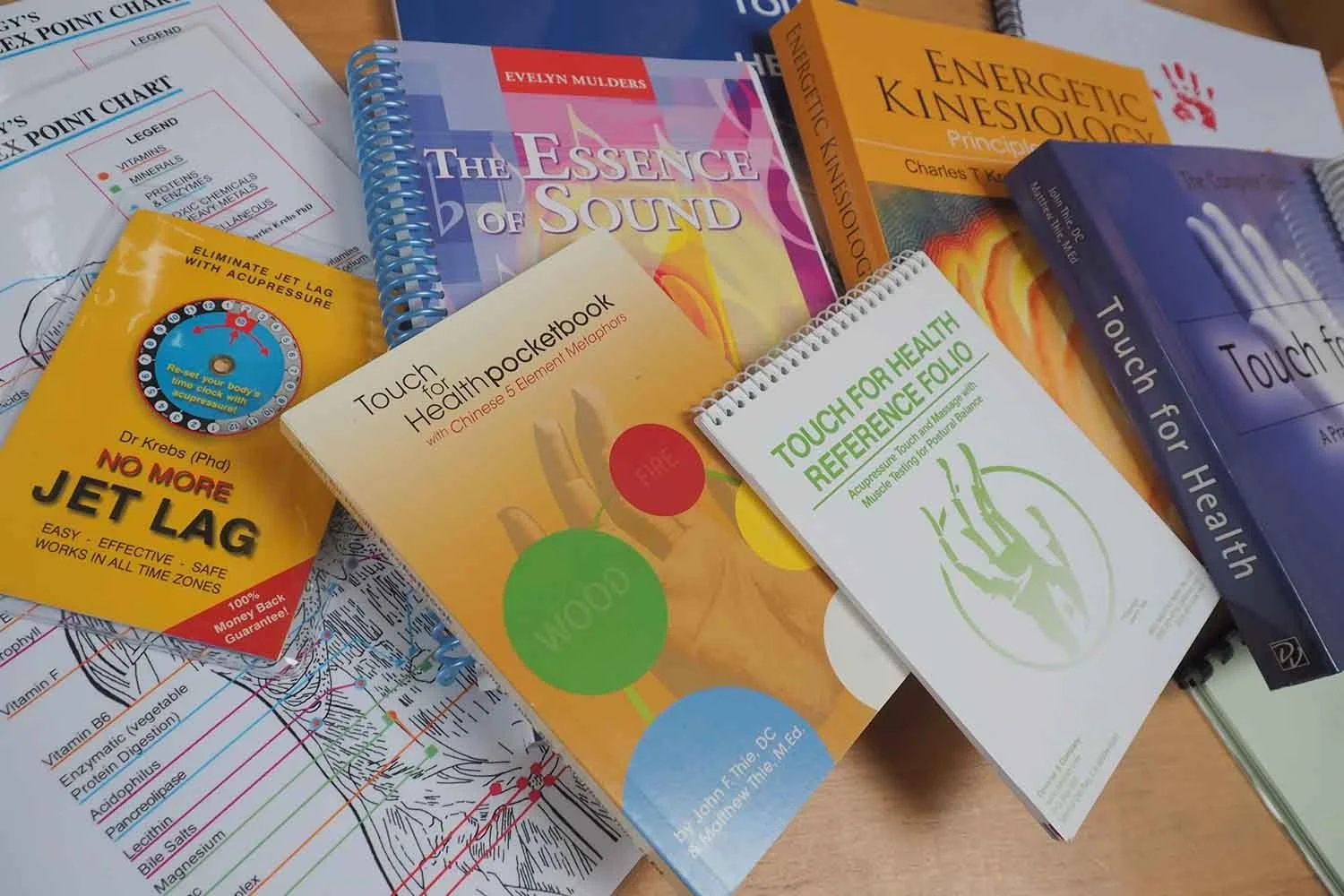 A collection of books and pamphlets on health and wellness, including titles like 'The Essence of Sound,' 'Energetic Kinesiology Principles,' 'Touch for Health,' and 'Touch for Health Reference Folio,' along with a diagram on a sheet of paper and a yellow card about jet lag remedies.