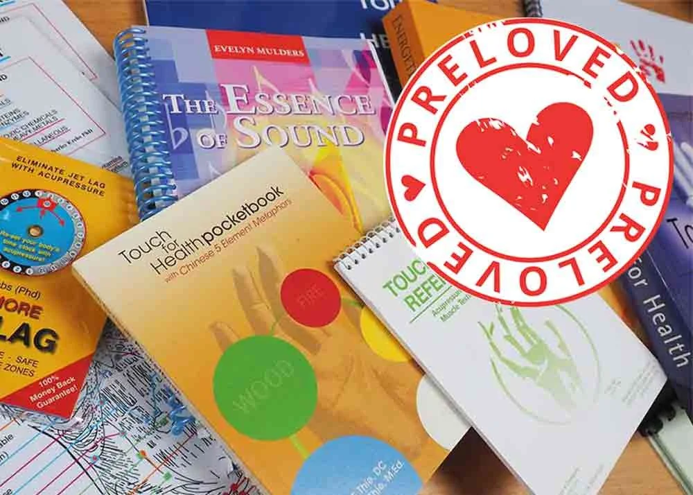 A collection of health and wellness books and pamphlets, including one titled 'Touch for Health Pocketbook,' with a red 'PRELOVED' sticker featuring a heart and a couple kissing, on a wooden surface.
