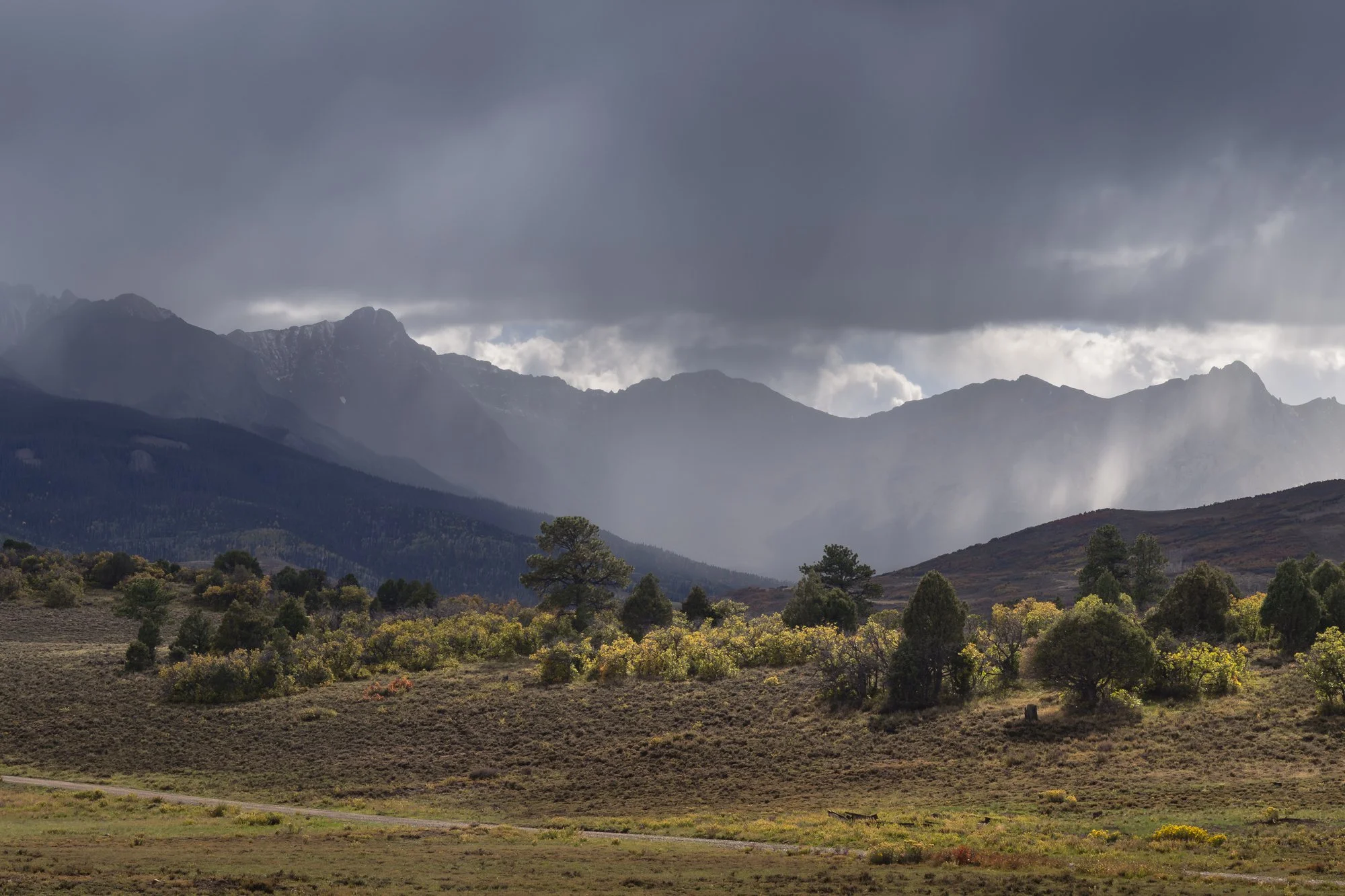 A mountainous landscape with dark clouds overhead and rays of sunlight breaking through, illuminating the valley and trees below.