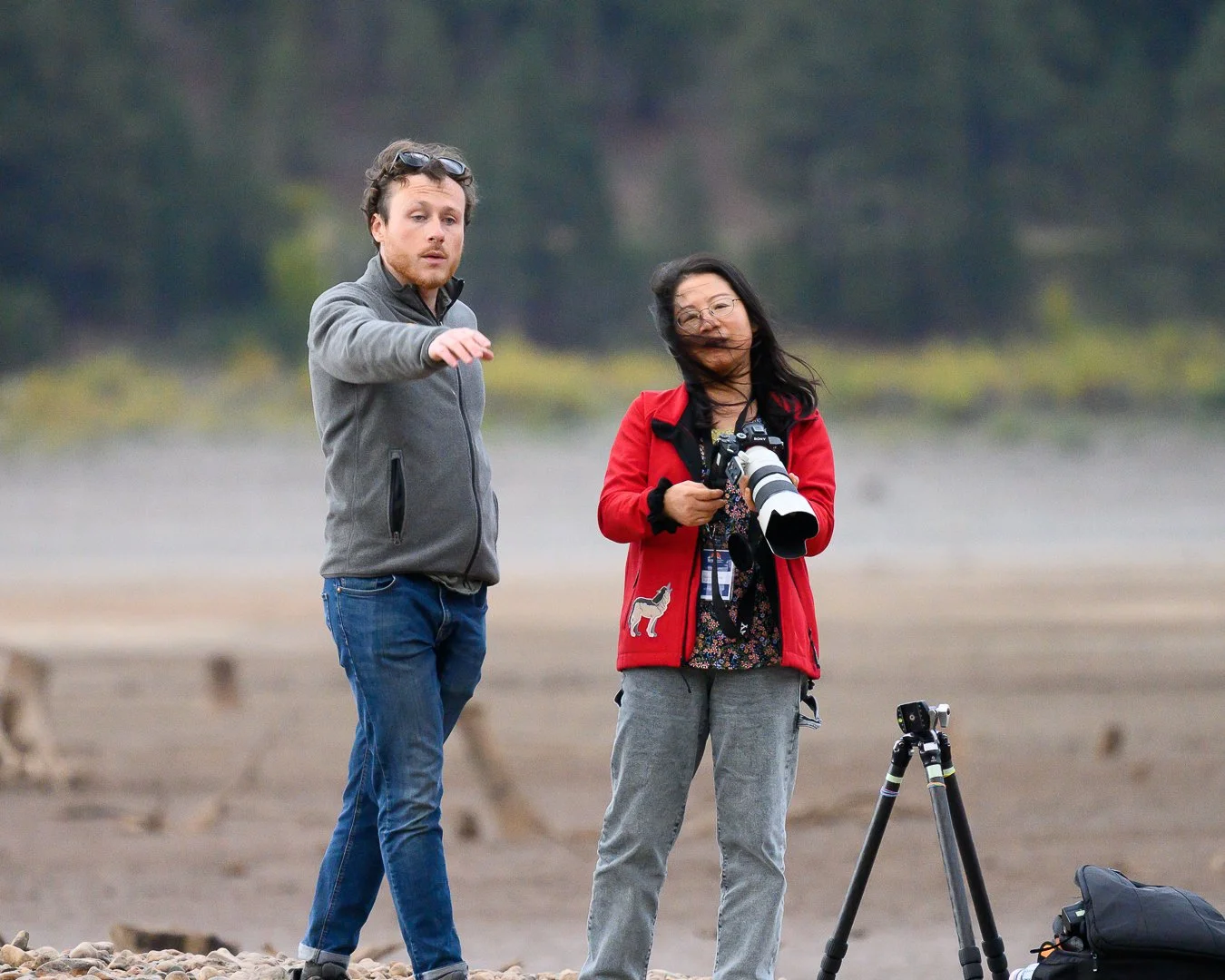 Two people are outside with photography equipment, one man and one woman, with the woman holding a camera and the man pointing at something in the distance. The woman is wearing a red jacket and glasses, and the man is in a gray jacket. There is a ca