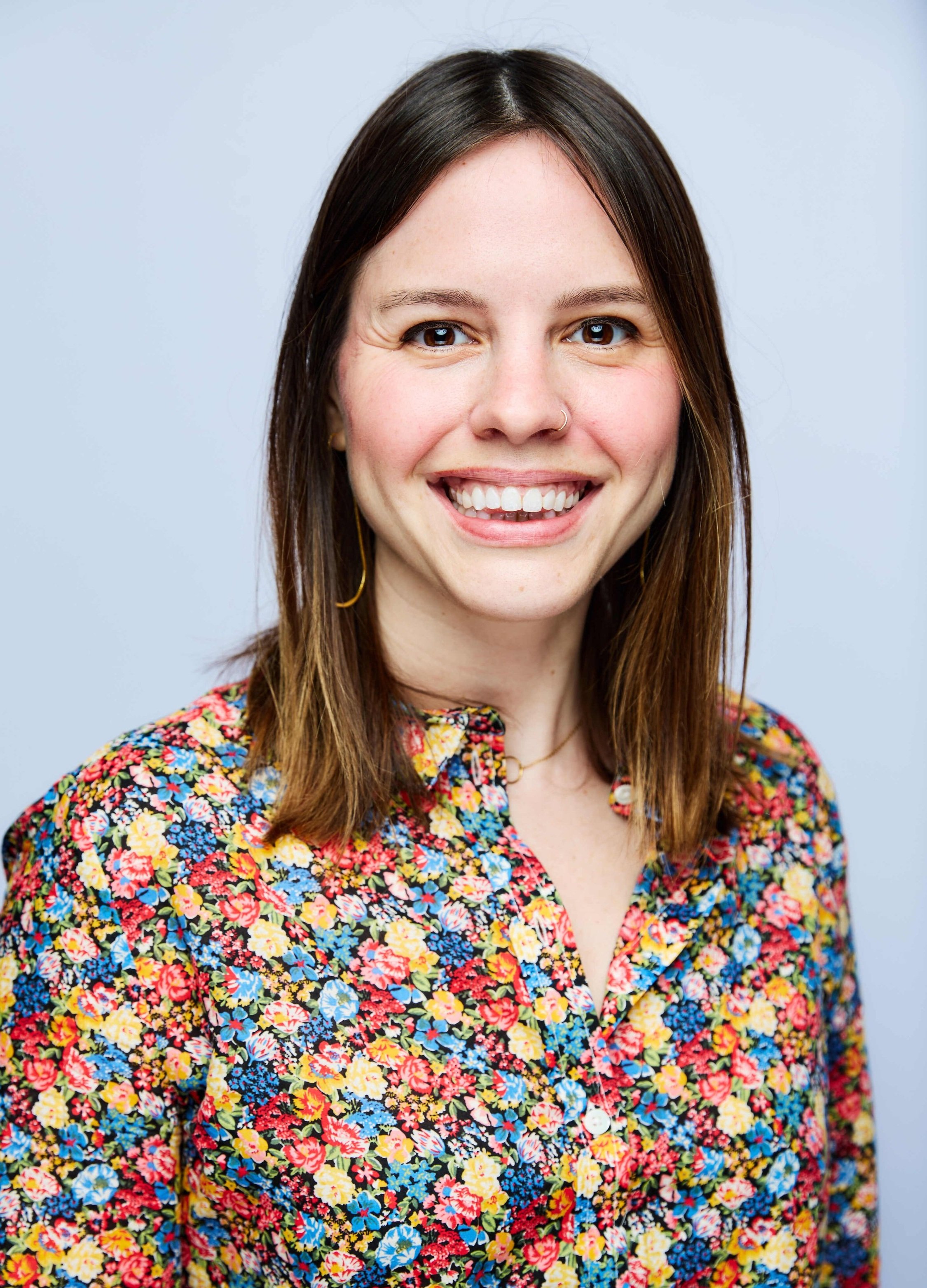 Portrait of a woman with long brown hair, wearing earrings, and a colorful floral blouse, smiling against a light blue background.