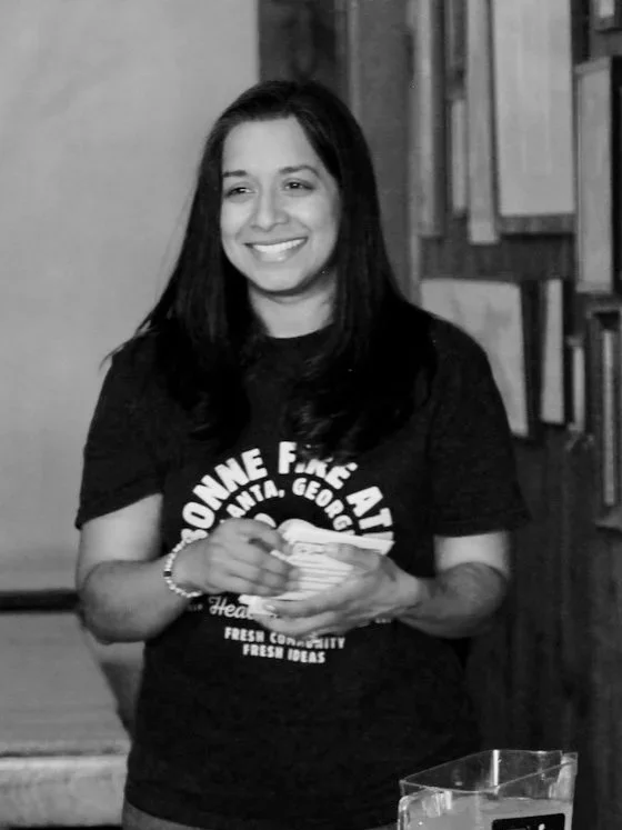 A woman with long dark hair smiling, holding a stack of papers or a napkin, standing indoors in front of a wooden wall and a table with a glass container.