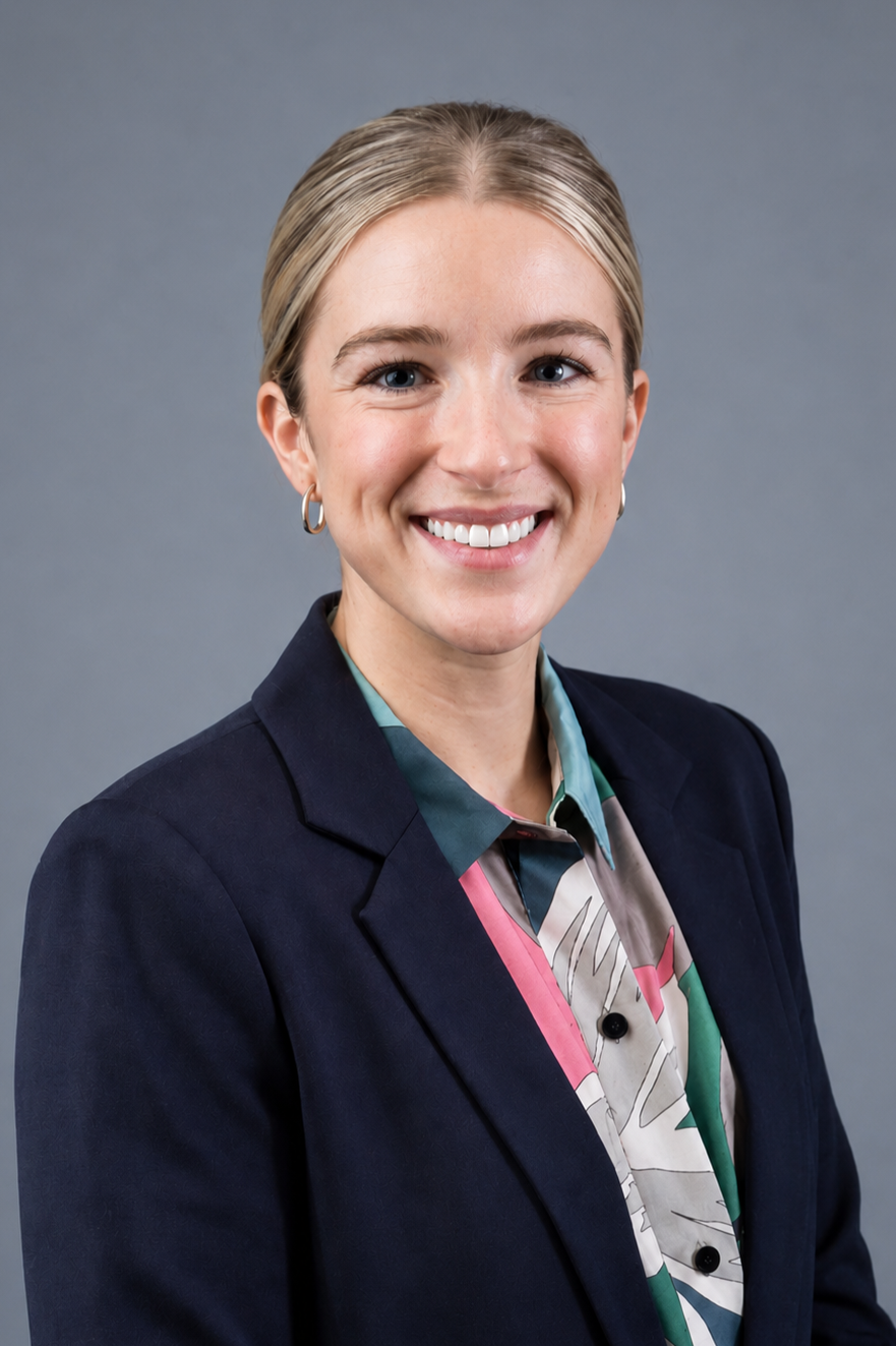 A woman with blonde hair in a bun, wearing a dark blazer over a colorful patterned blouse, smiling at the camera against a gray background.