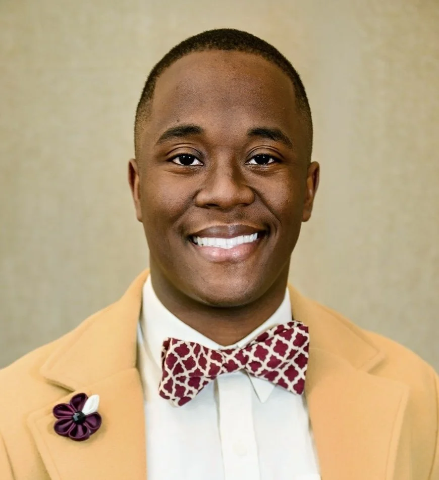 A smiling man wearing a beige blazer, a white shirt, and a red patterned bow tie.