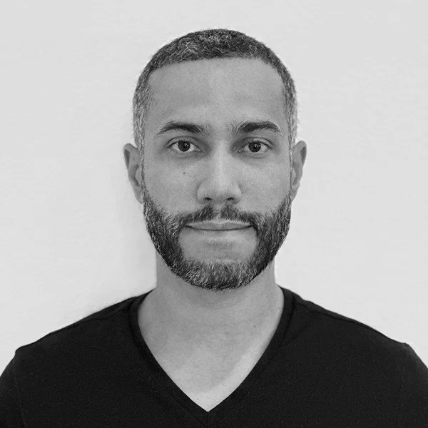 Portrait of a man with short hair and a beard, wearing a black V-neck shirt, standing against a plain white background.