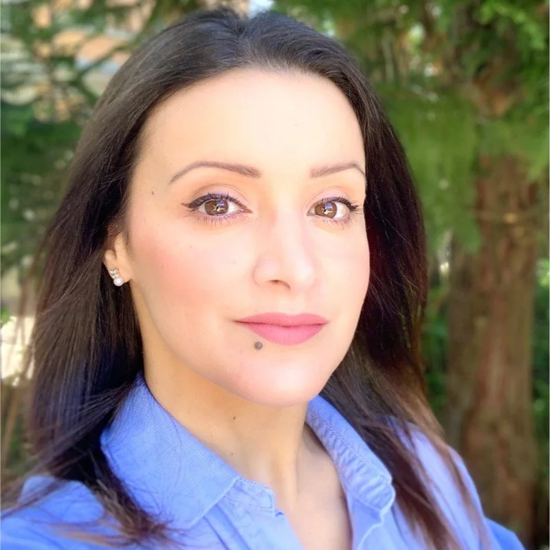 Close-up portrait of a woman with fair skin, dark brown hair, and natural makeup, wearing a blue collared shirt, standing outdoors with green trees in the background.