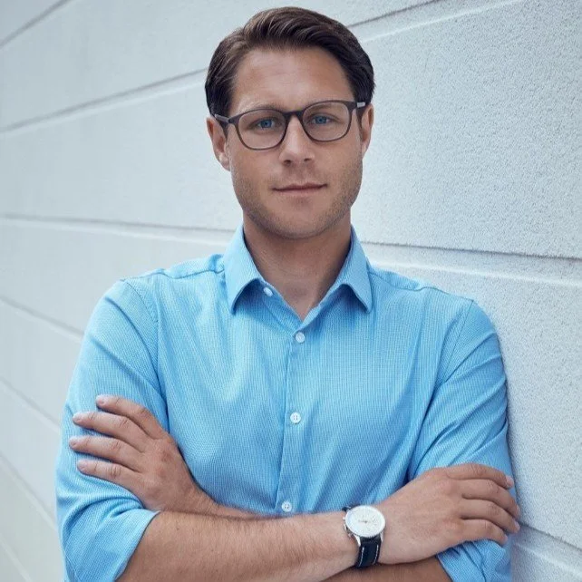 A young man with brown hair, wearing black-rimmed glasses and a light blue button-up shirt, standing against a white wall with his arms crossed.