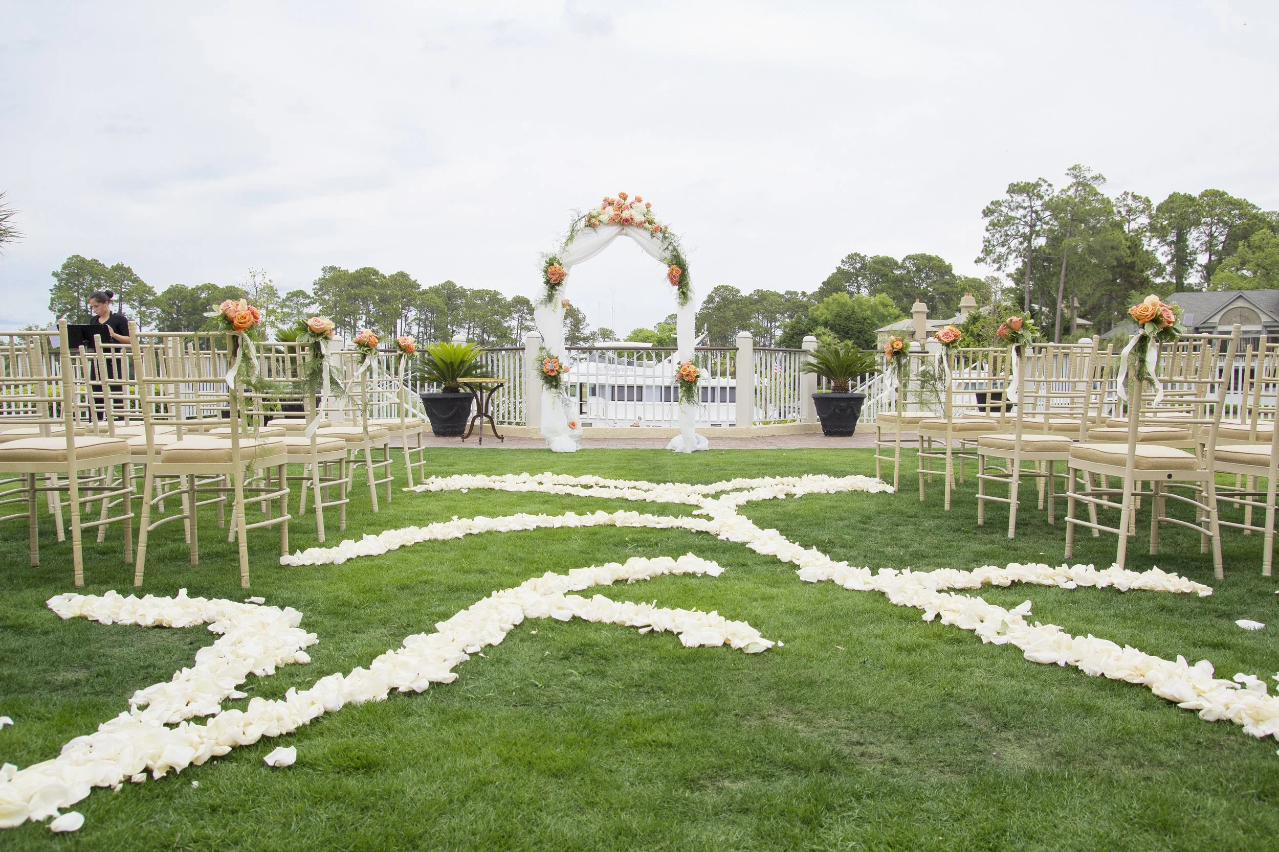 Wedding ceremony setup on a grass lawn with chairs decorated with pink roses, a floral arch, and petal pathways.