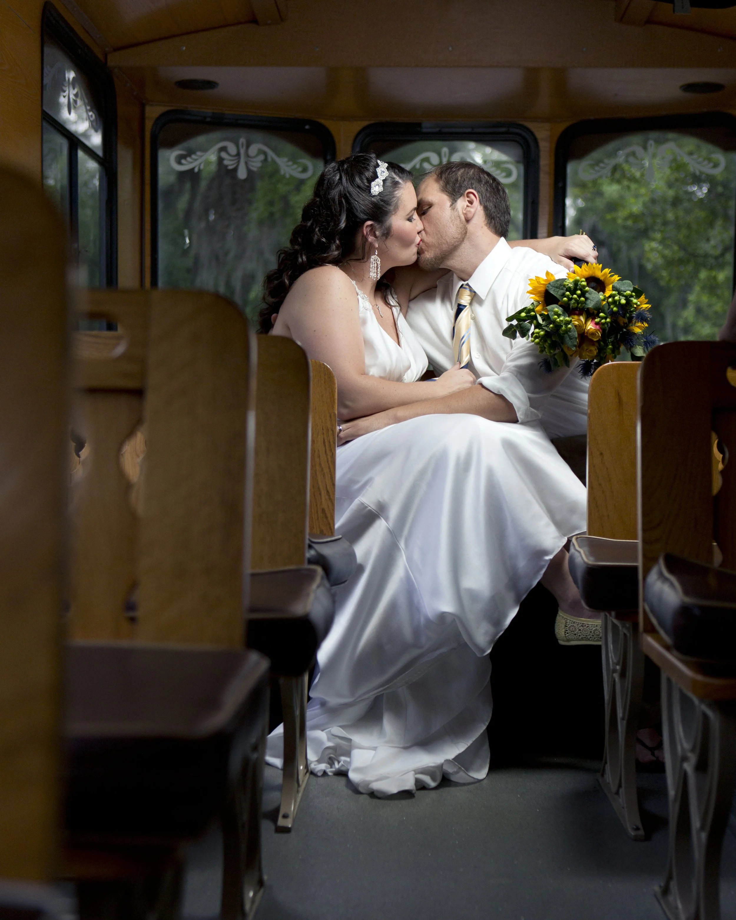 A bride and groom sit inside a bus, sharing a kiss. The bride is in a white wedding dress with dark, curly hair and a headband. The groom is in a white shirt with rolled-up sleeves and a striped tie, holding a bouquet of yellow and orange flowers. Th