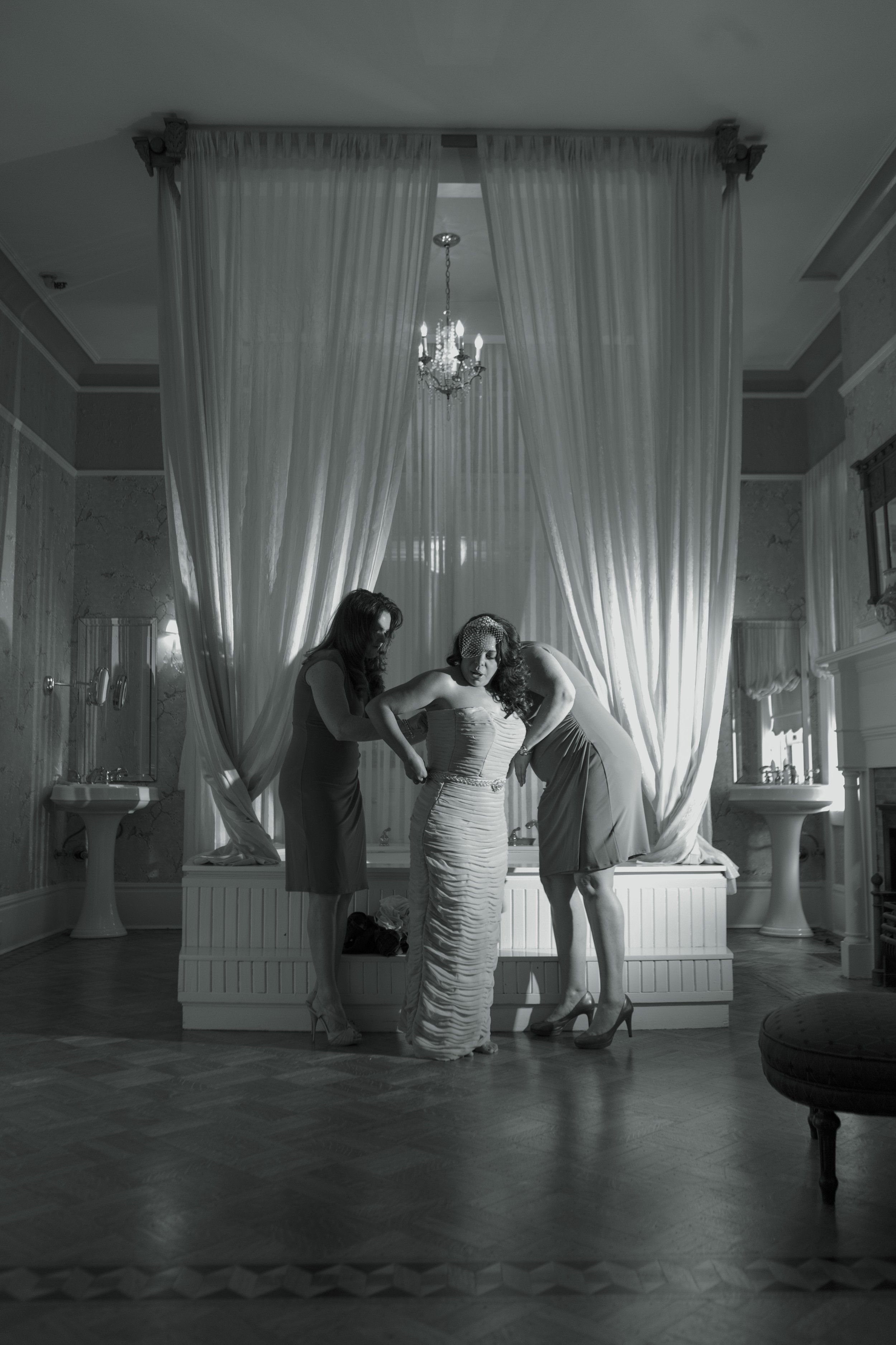 Three women in formal dresses, one in a wedding gown, standing in a bedroom with draped curtains, chandeliers, and sinks, adjusting the bride's dress.