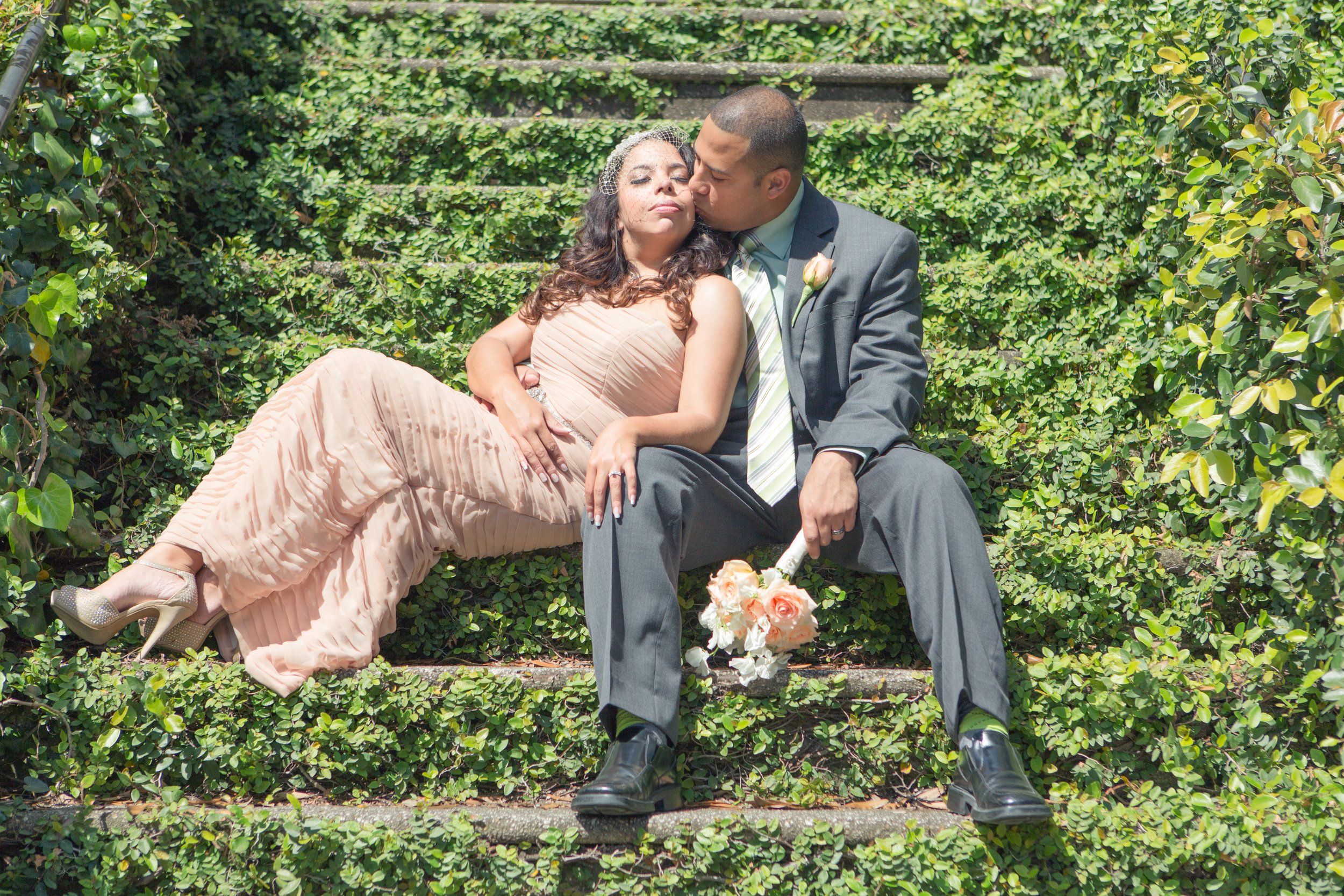 A couple in formal attire sitting on garden stairs covered with green leaves; the woman is resting against the man with her eyes closed, and he is kissing her on the forehead.