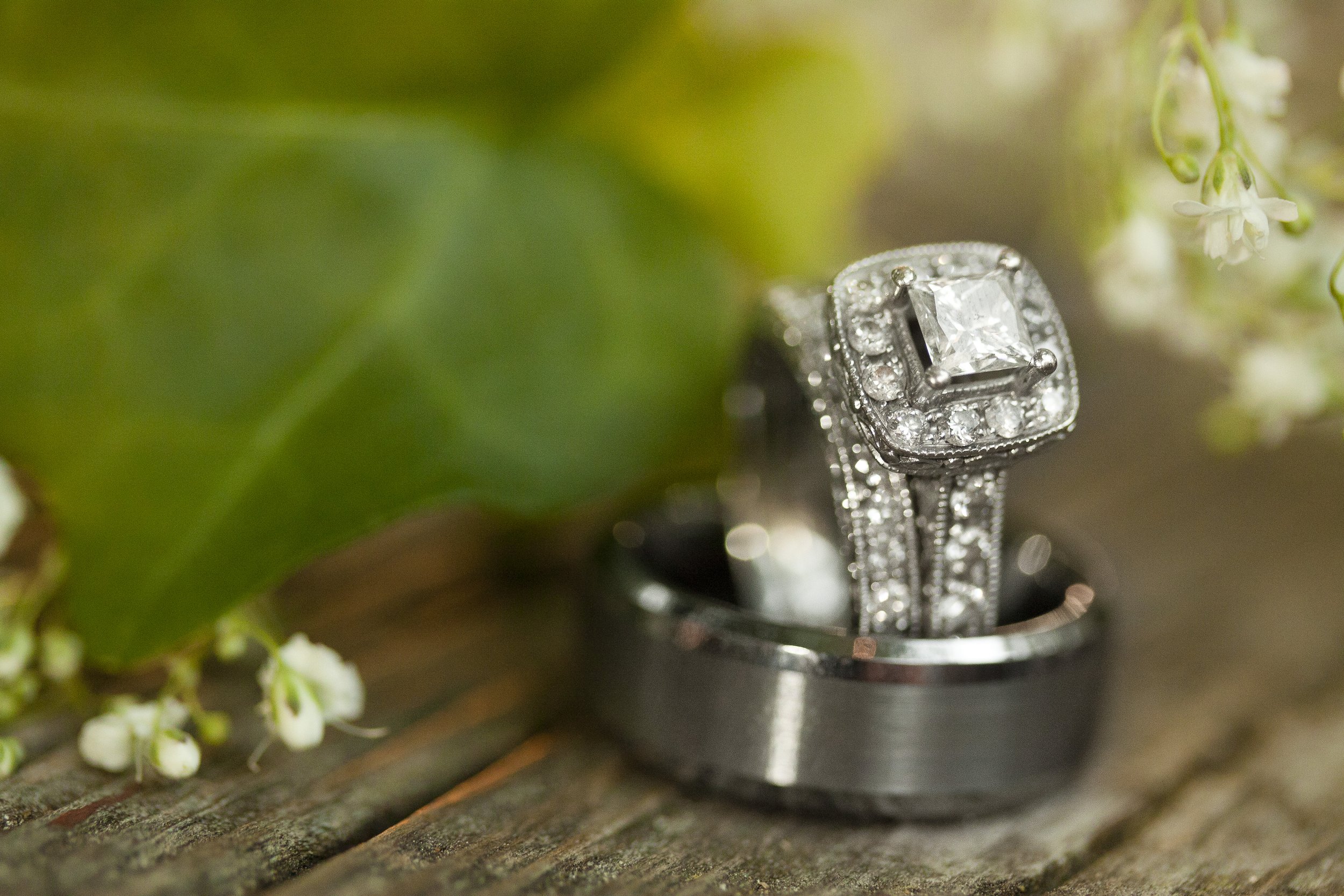 Close-up of a engagement ring with a square diamond in the center, surrounded by smaller diamonds, placed on a wooden surface next to a wedding band, with green leaves and small white flowers nearby.