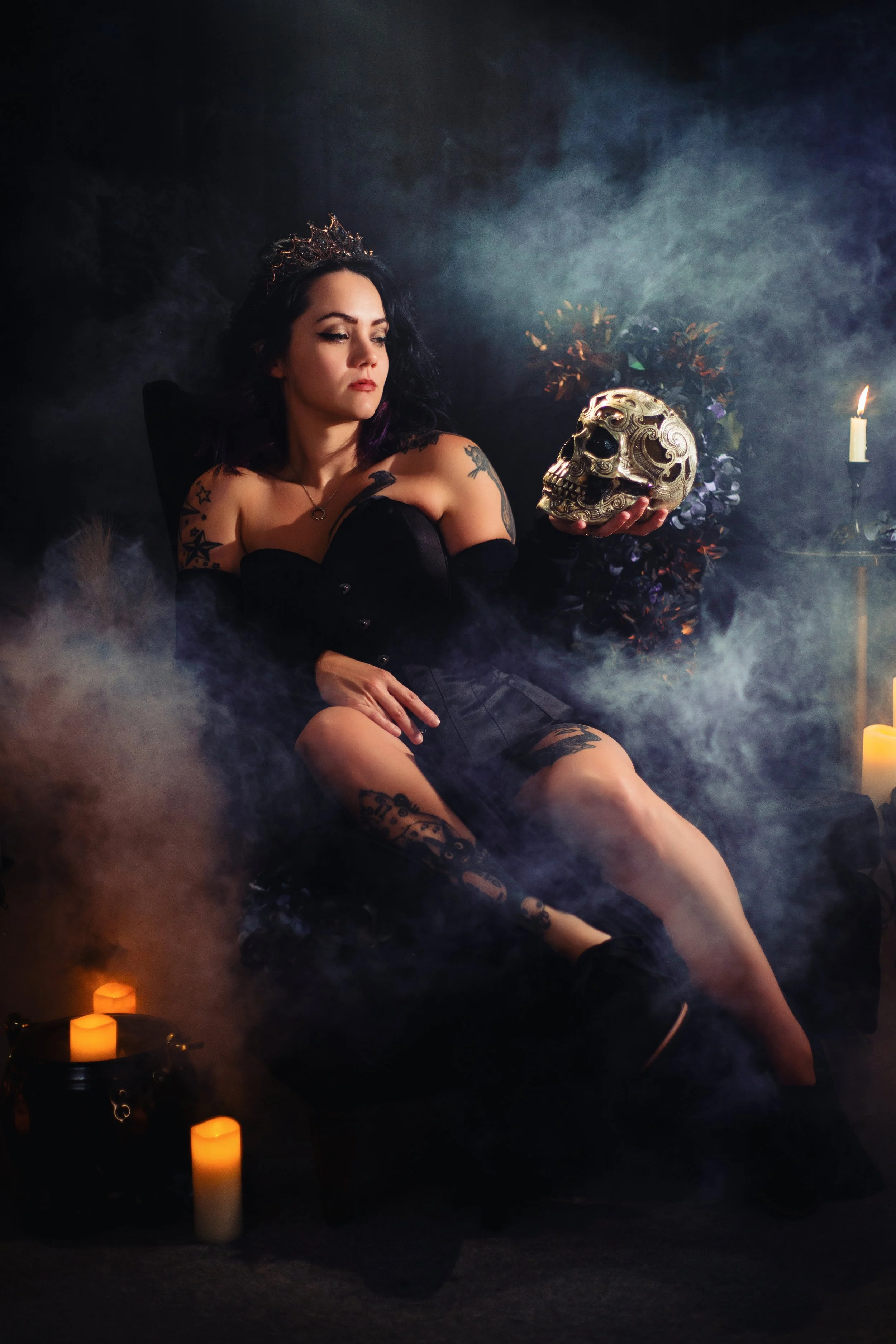 A woman dressed in black with tattoos, sitting in a dark, smoky room, holding a decorative skull and surrounded by lit candles.