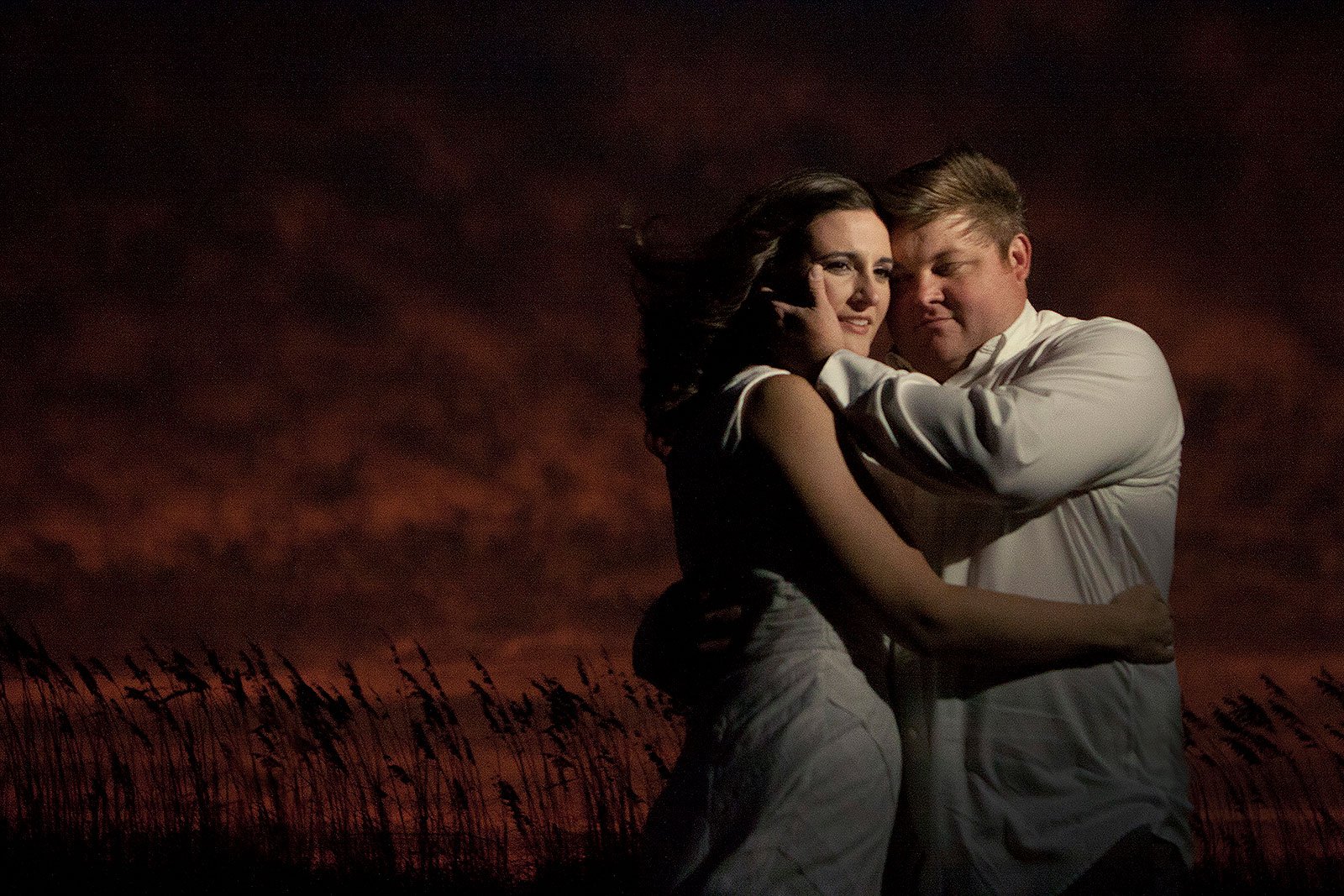 A man and woman embrace each other outdoors at sunset or dusk, with a dark sky and tall grasses visible in the background.