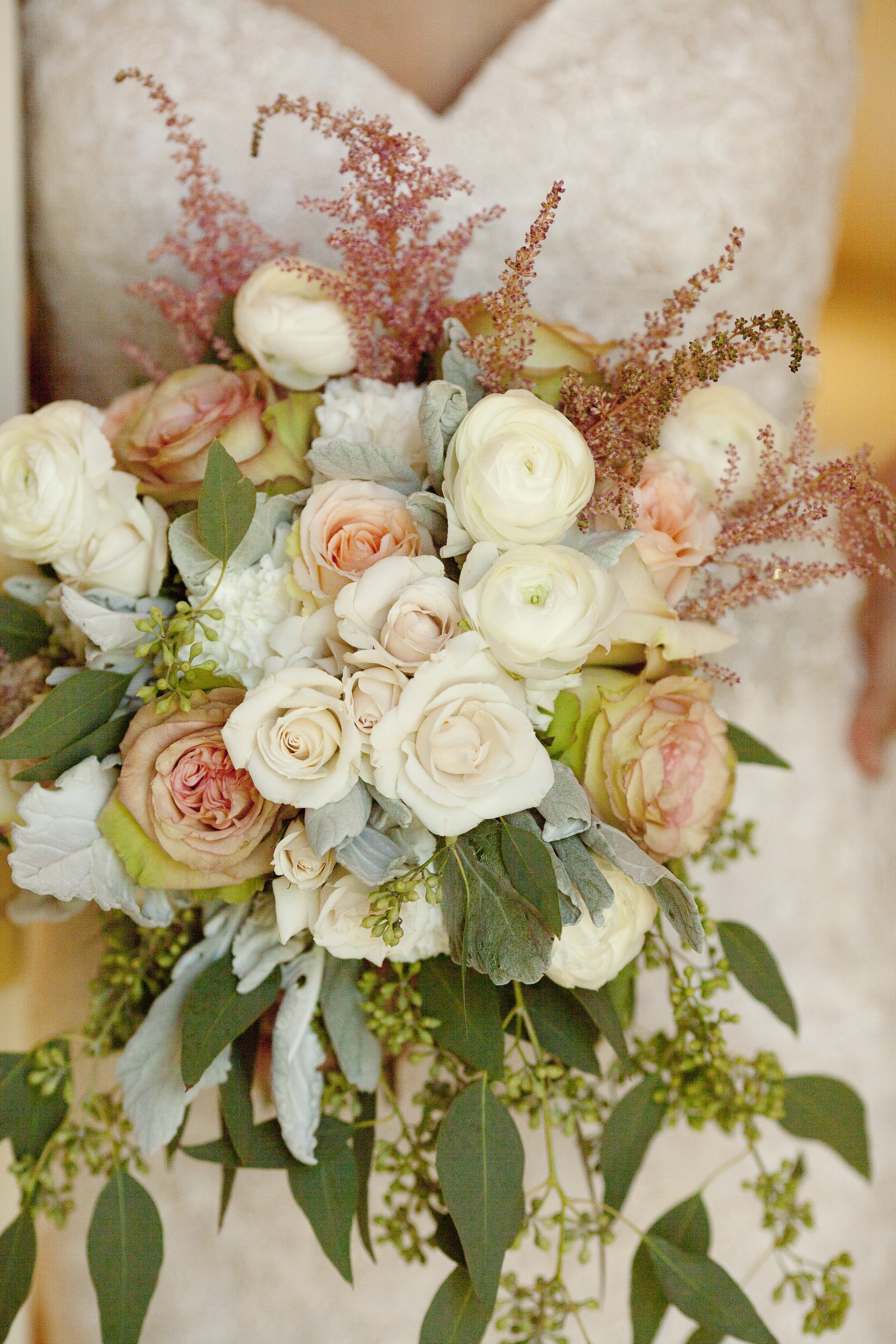 A bridal bouquet with white and blush roses, pink Astilbe, and green foliage held by a person in a white lace dress.