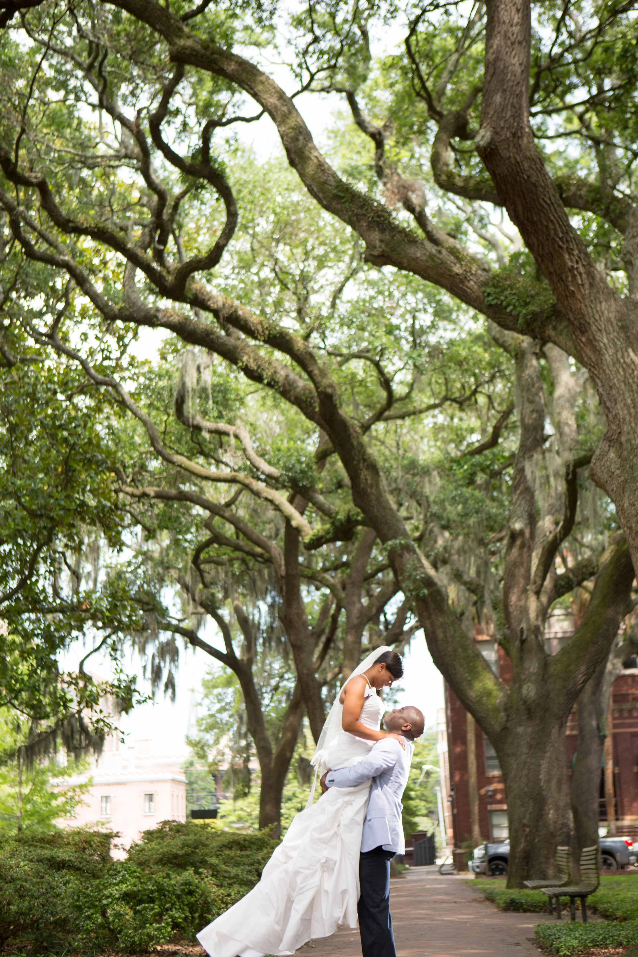 A bride in a white wedding dress being lifted by a groom in a light gray suit outdoors, surrounded by large trees and greenery.