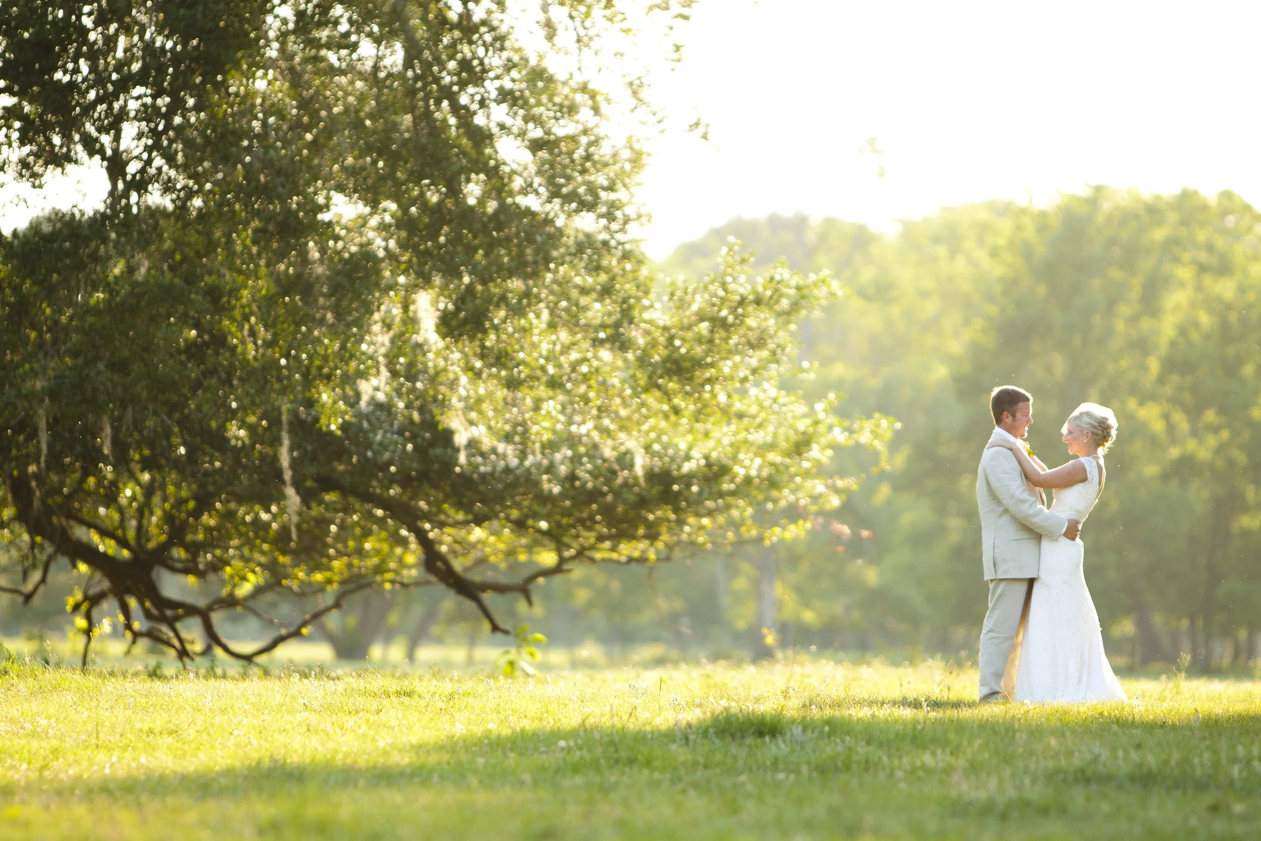A couple dressed in wedding attire standing on a grassy field, holding each other and smiling under large trees with sunlight streaming through the leaves in the background.
