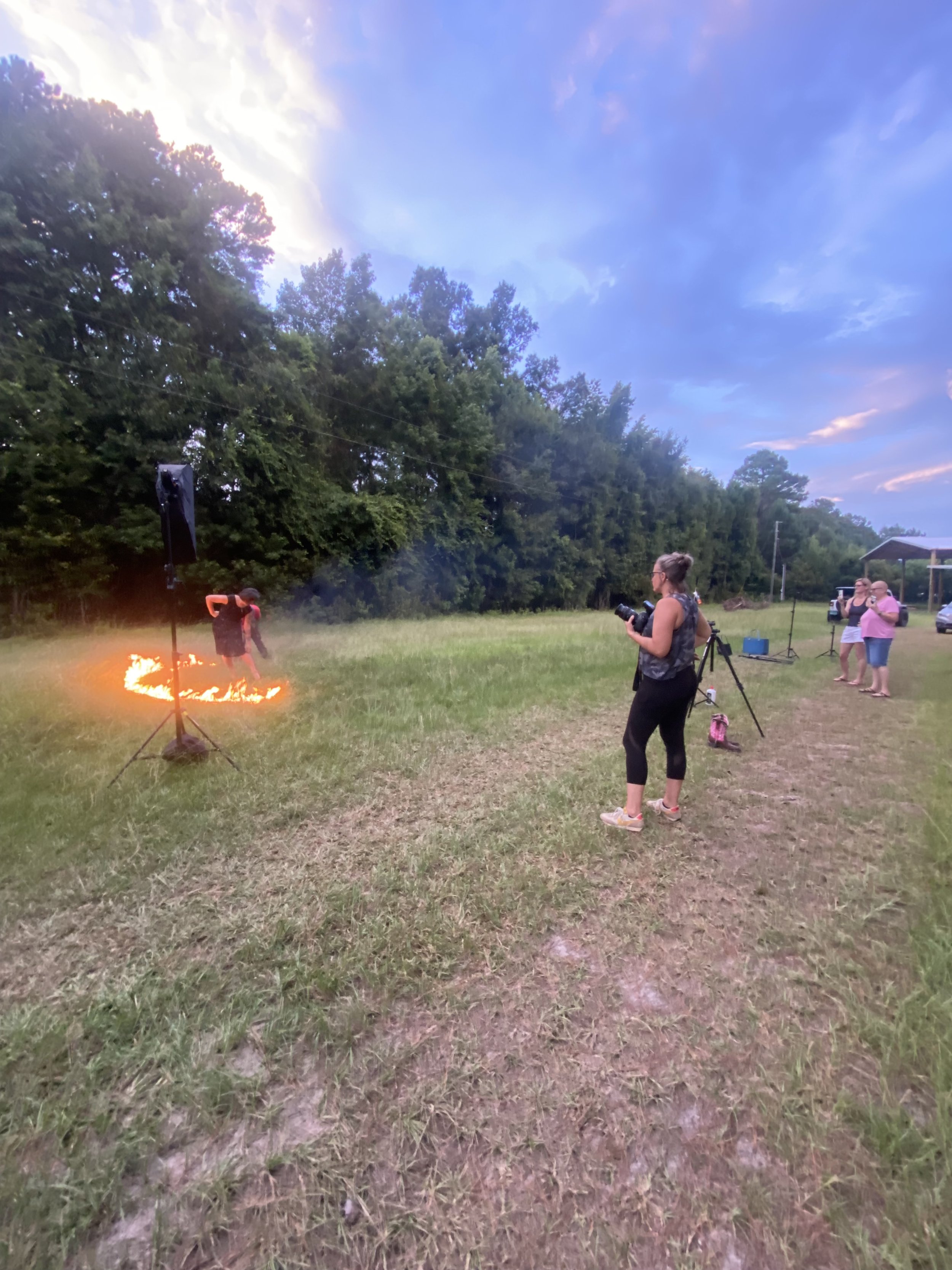 A fire performer creating a flame circle on the grass at dusk, with three women observing and a woman taking a photo, in an open field surrounded by trees.