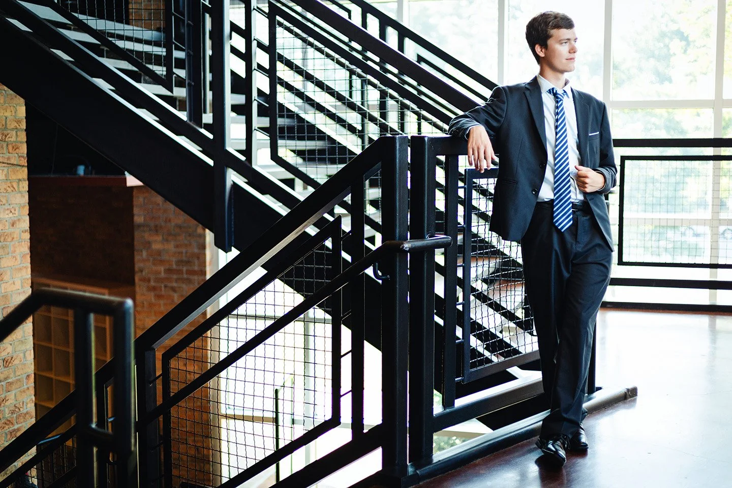 A young man in a business suit leaning on a metal railing in a modern office building with large windows and natural light.