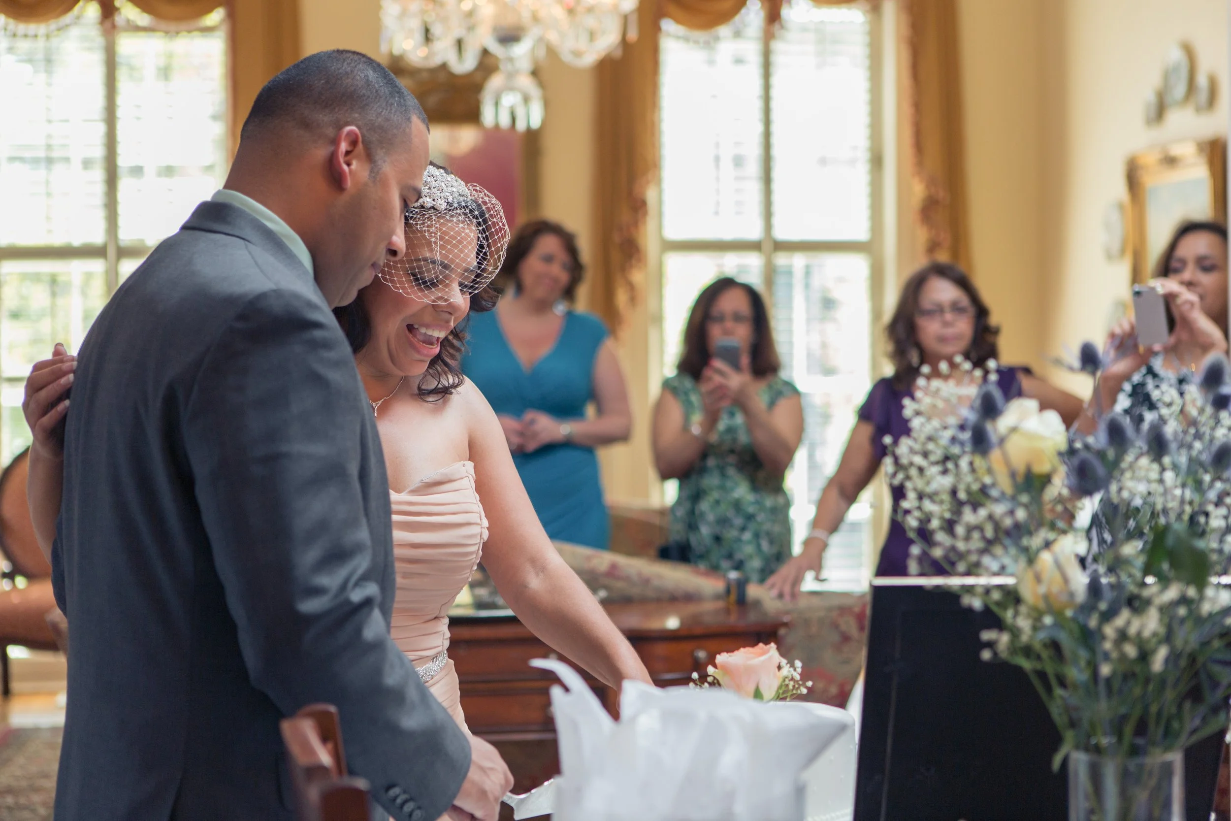 A bride and groom sharing a joyful moment, surrounded by friends taking photos, with a decorated bouquet on a table indoors with large windows and elegant curtains.