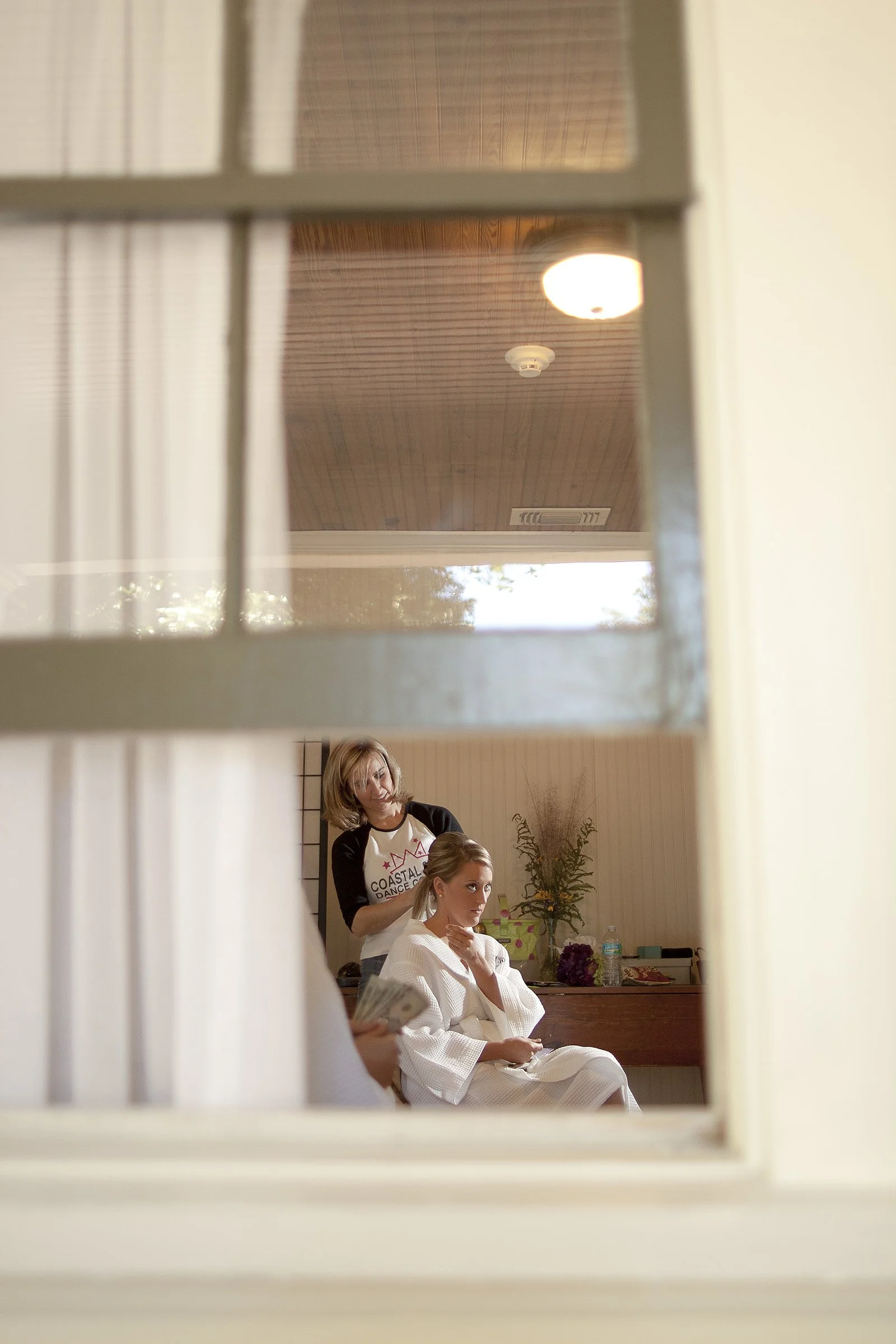 A woman is sitting in a room wearing a white robe, while a hairstylist stands behind her doing her hair. The image is viewed through a window with grid panes.