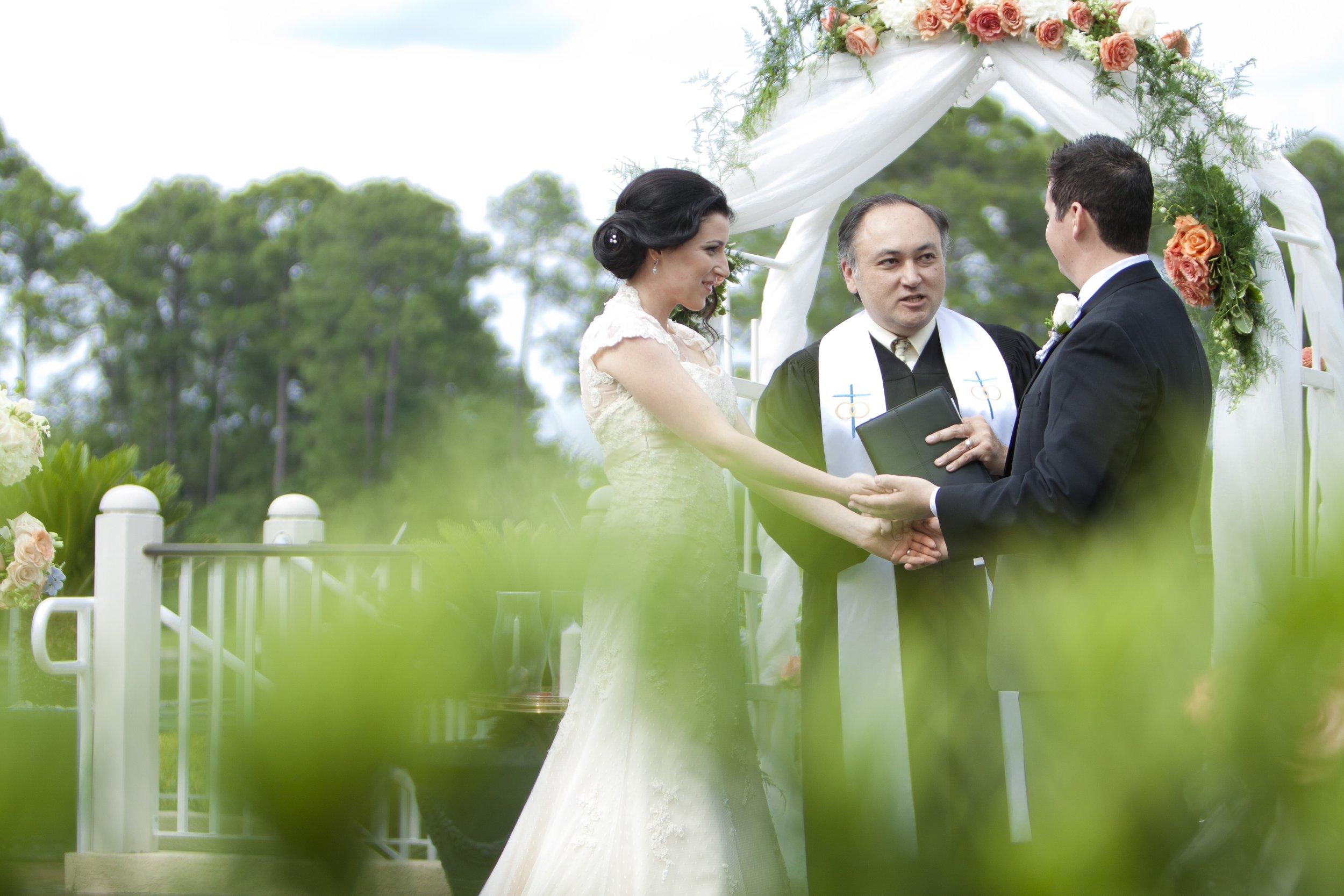 A couple getting married at an outdoor wedding ceremony, with a minister officiating, under a floral arch with green trees in the background.