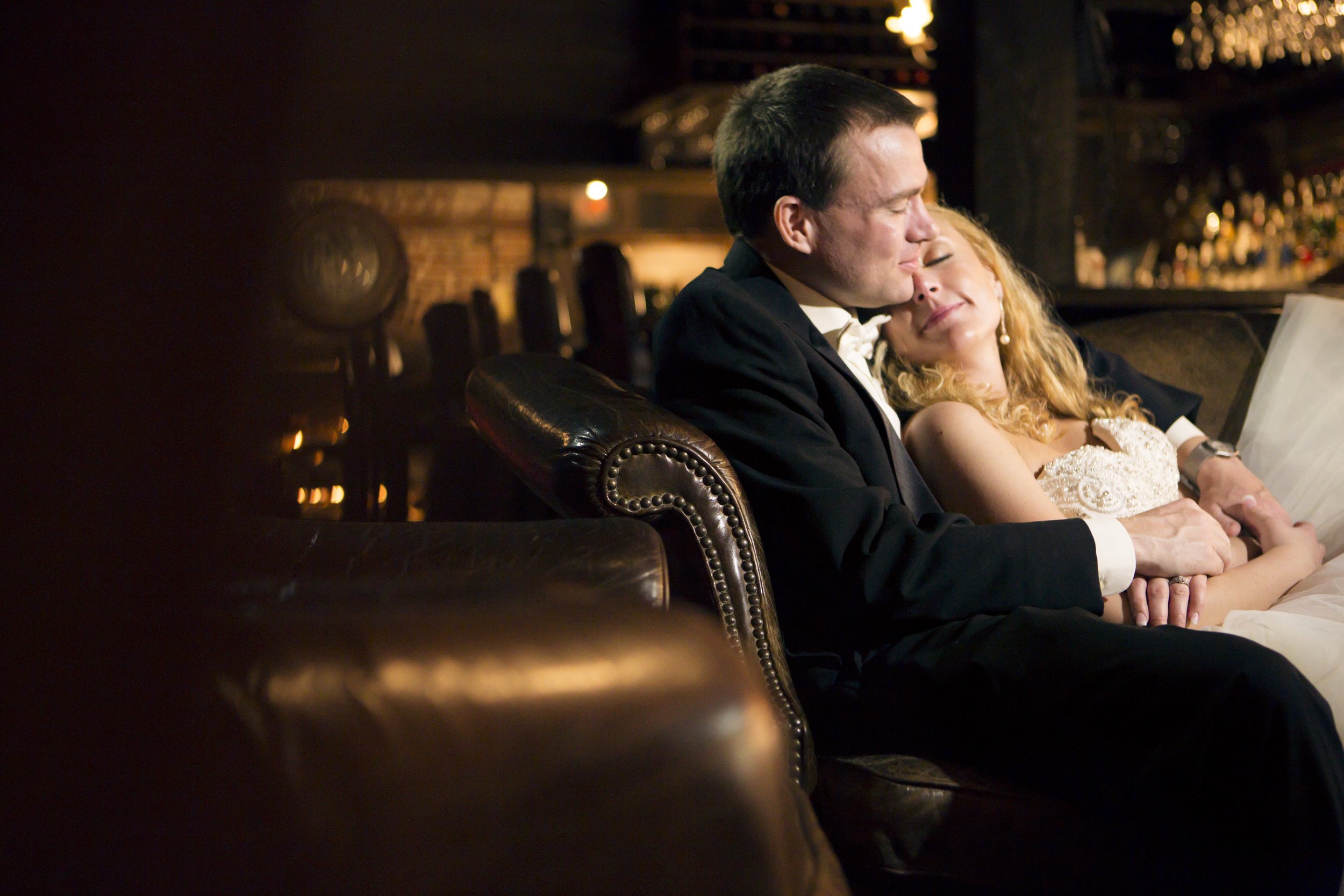 A man in a tuxedo and a woman in a white dress are sitting close together on a leather couch, with their eyes closed and faces touching, in a warmly lit restaurant or bar setting.