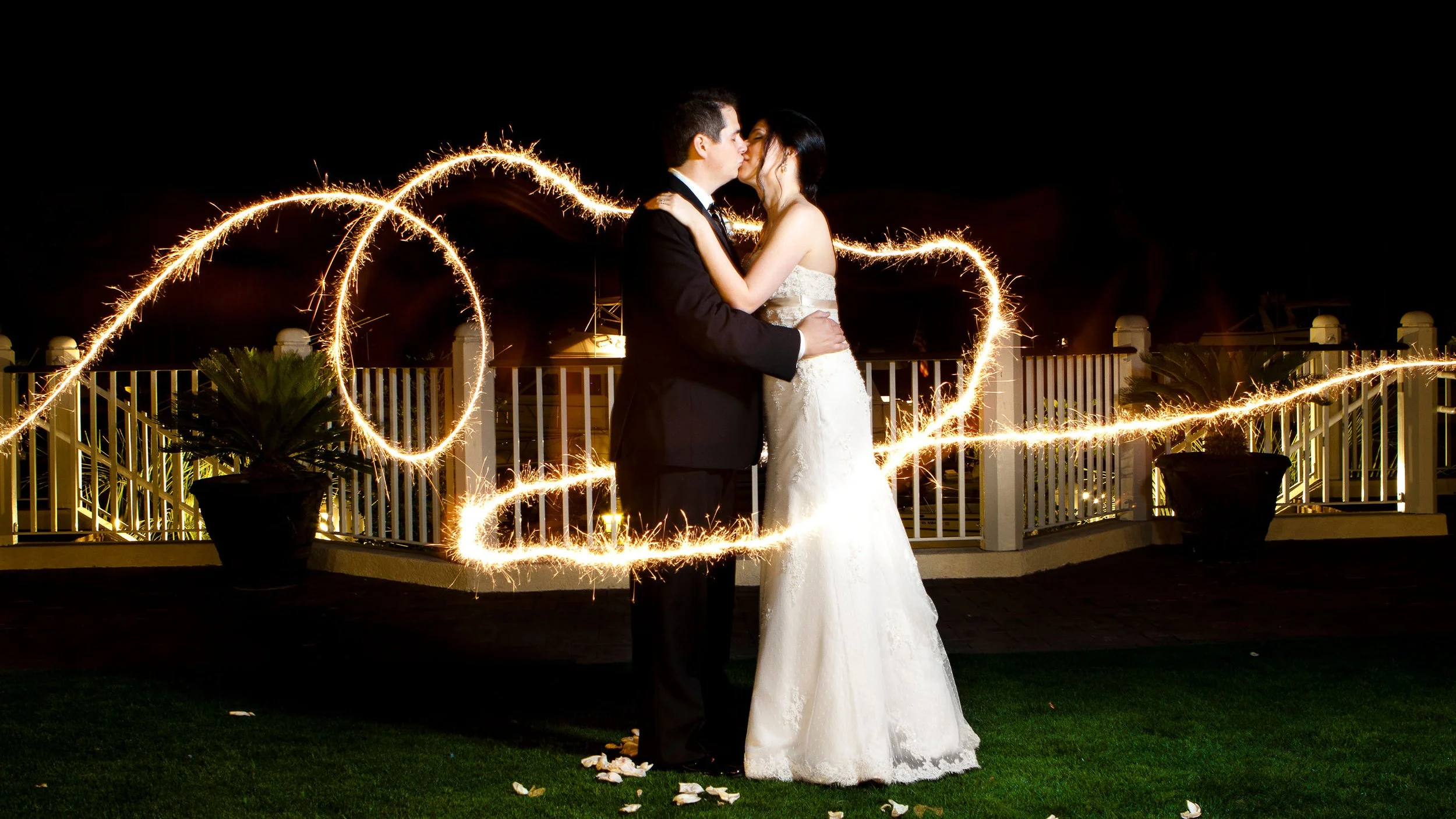 A bride and groom kiss on their wedding night, surrounded by sparklers forming the number 2023, on an outdoor balcony with potted plants and a white fence.