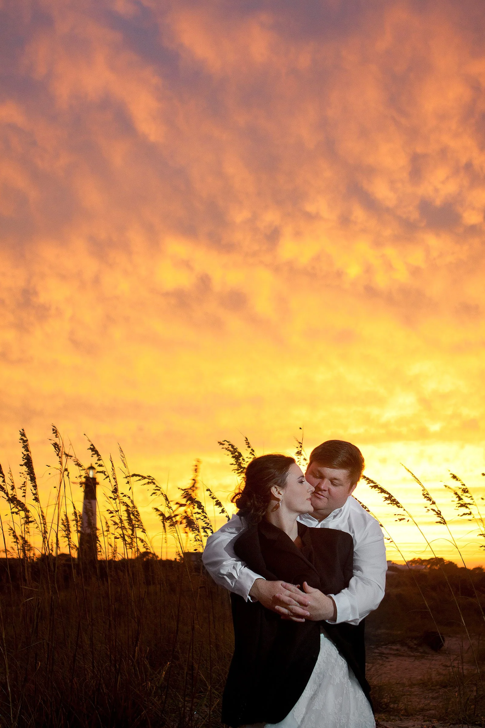 A couple dressed in wedding attire embracing outdoors during a sunset with a vibrant orange sky and tall grass in the background.