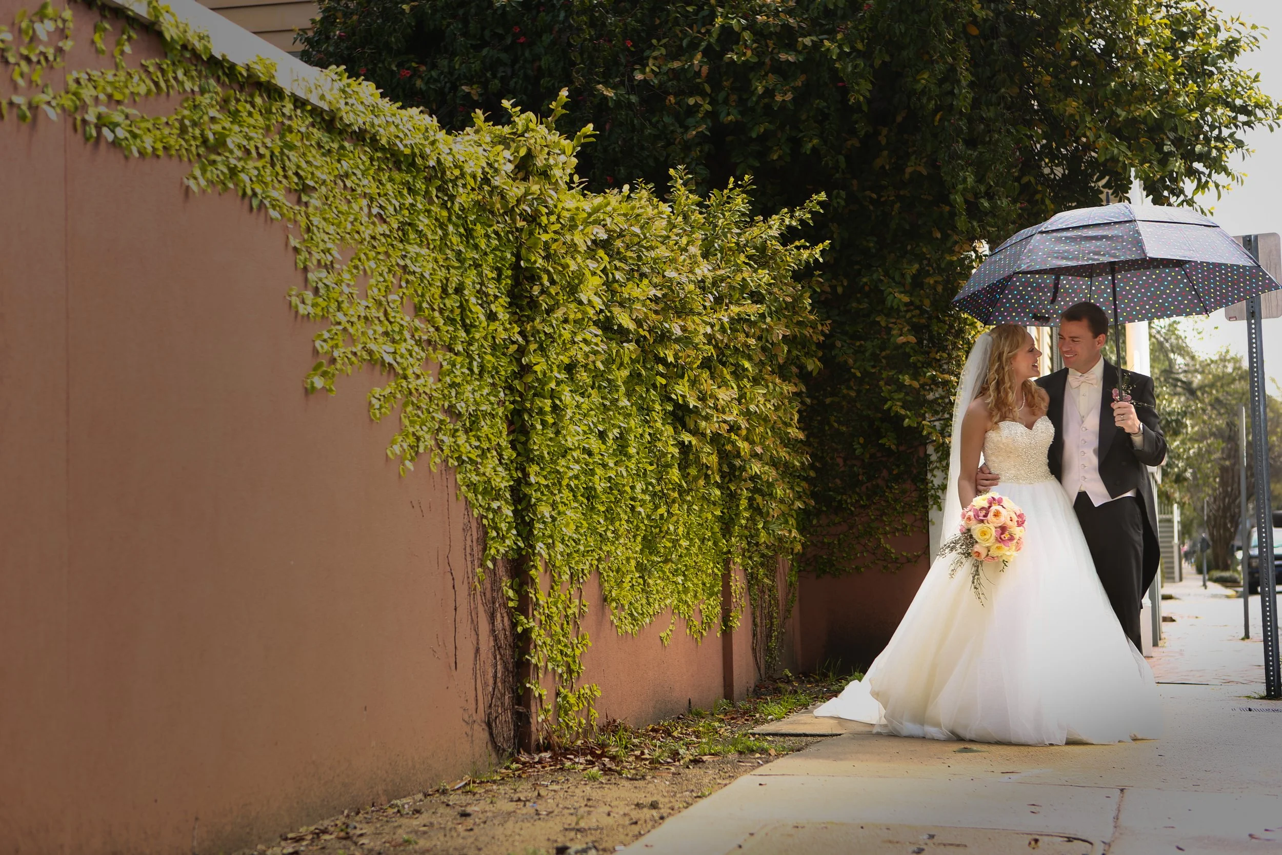 A newlywed couple walking on a city sidewalk under a large umbrella. The bride is in a white wedding dress holding a bouquet of flowers. The groom is dressed in a black tuxedo with a white bow tie. They are smiling and looking at each other, sharing 