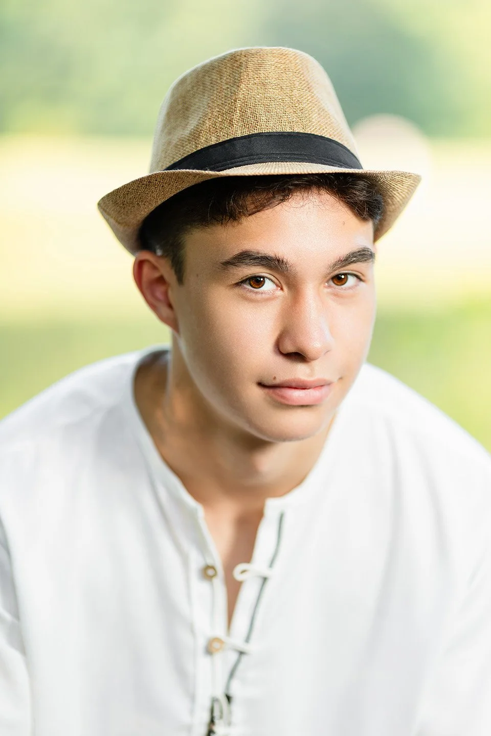 A young man wearing a beige fedora hat and a white henley shirt, outdoors with a blurred green background.