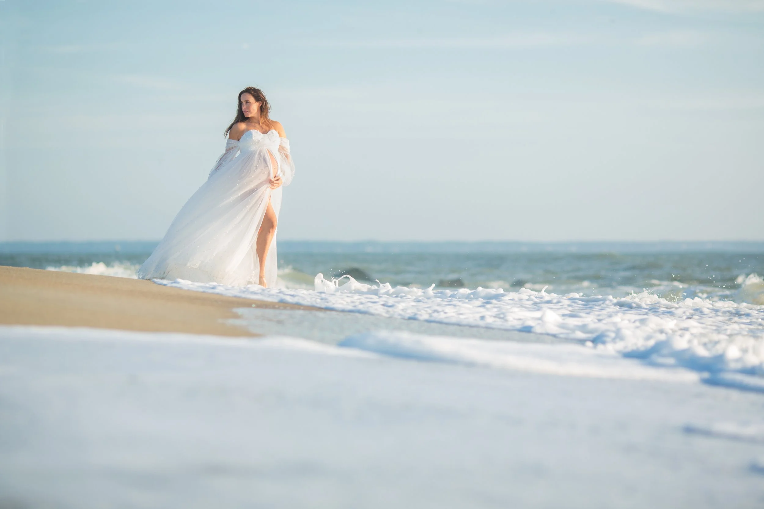 Woman in a white dress standing at the edge of the ocean on the beach.