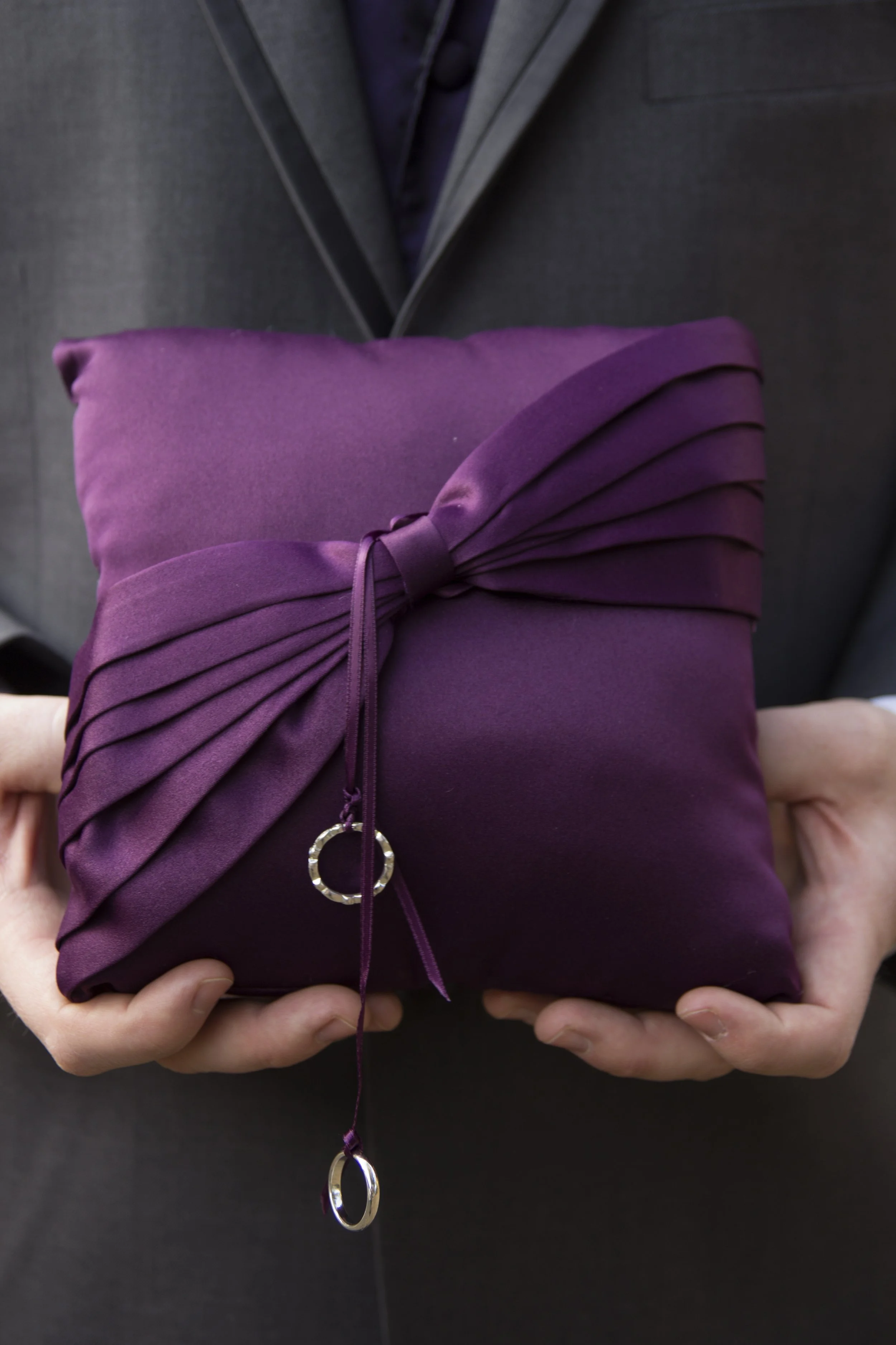 Person in dark suit holding a purple fabric gift box with pleated ribbon decoration and a jewelry charm.