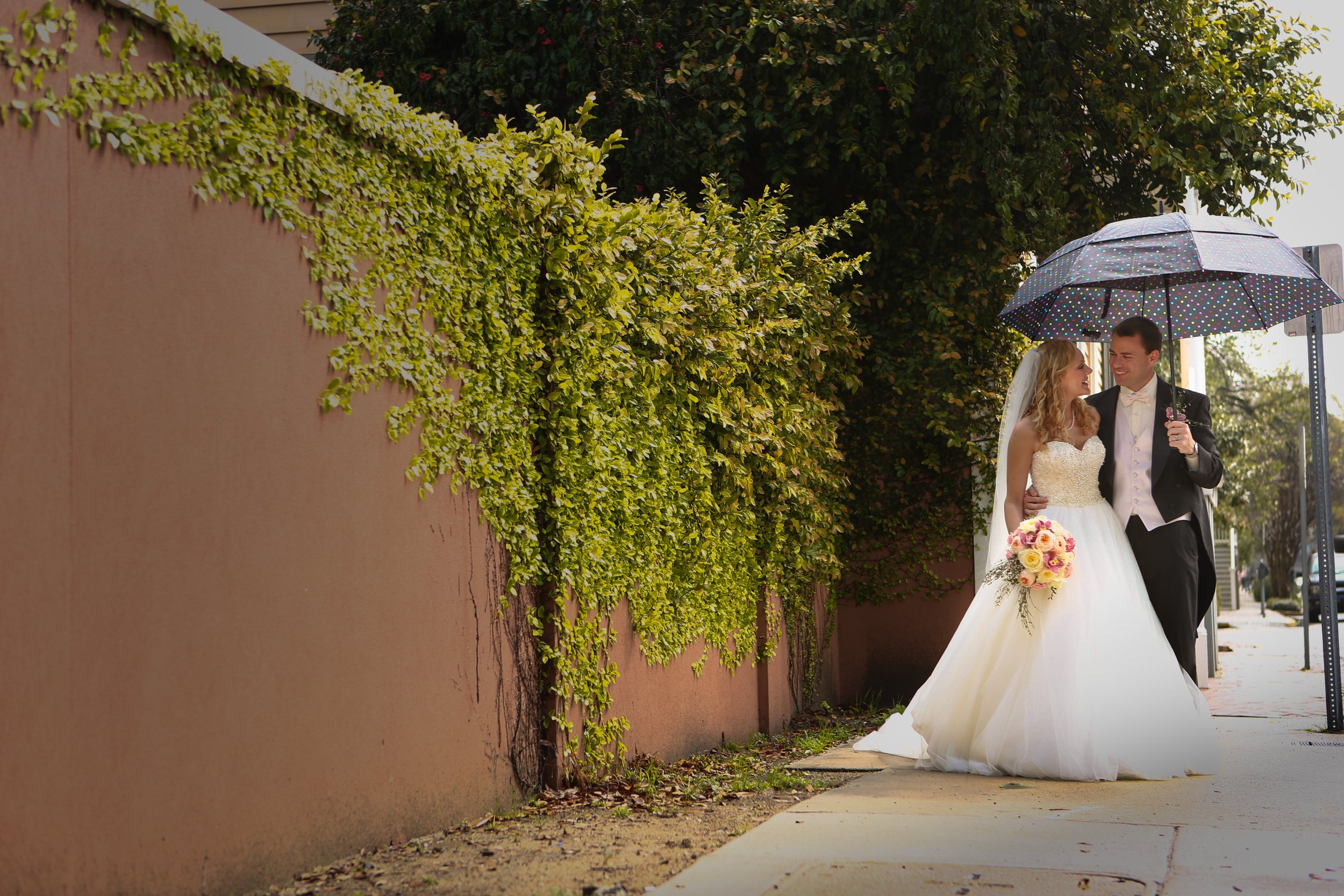 A bride and groom walking on a sidewalk under an umbrella, smiling and looking at each other, with greenery and a pink wall in the background.