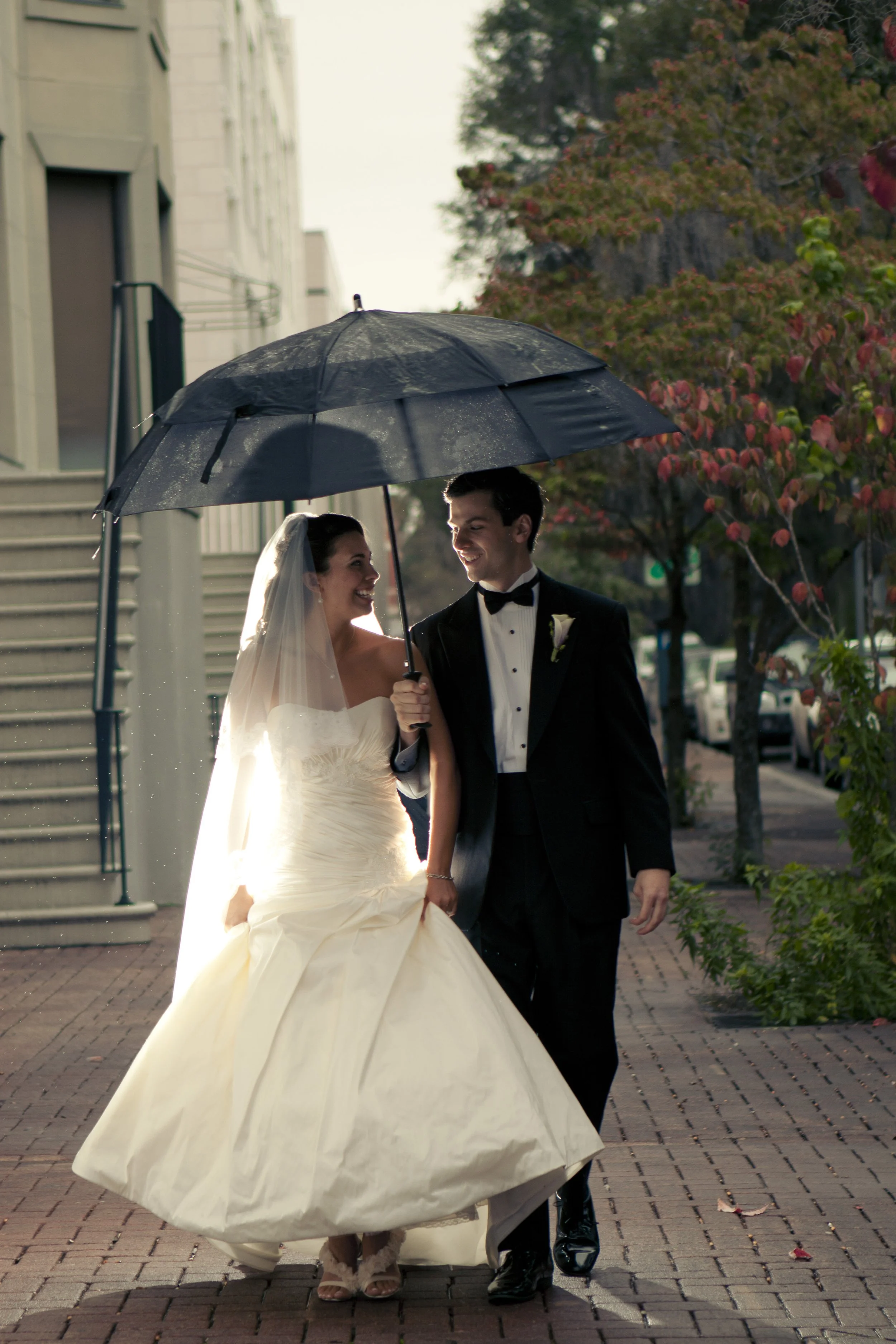Bride and groom walking outdoors in the rain under a large umbrella, smiling at each other.