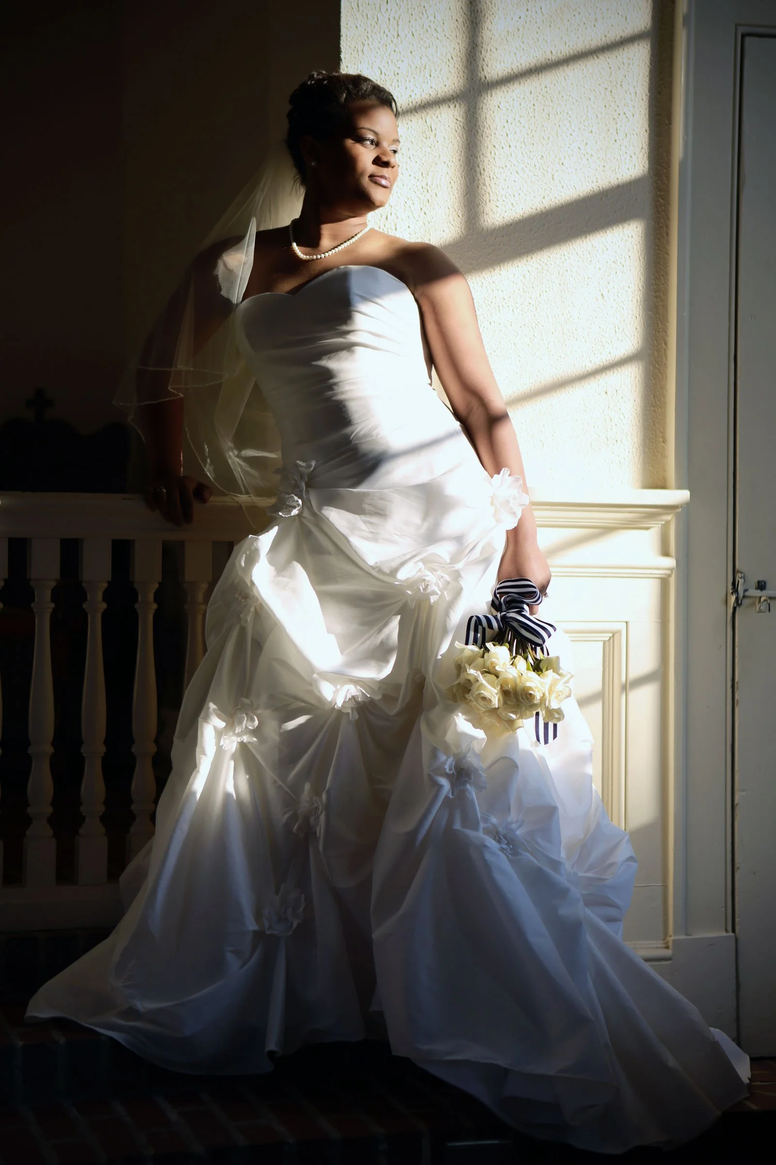 A bride in a strapless white wedding gown holding a bouquet of white roses with striped ribbons, standing by a window with sunlight casting shadows on the wall behind her.