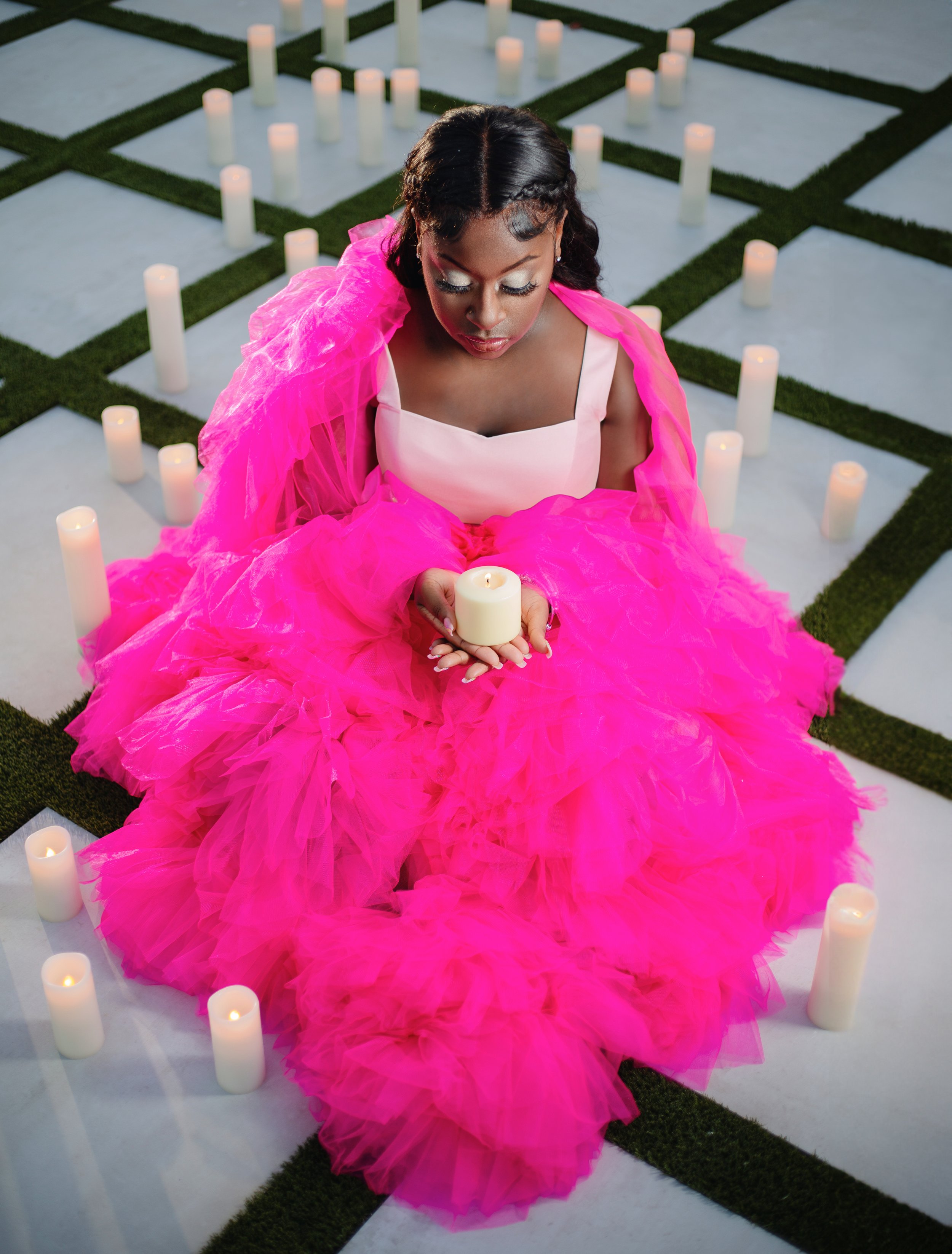 A woman in a bright pink gown sits cross-legged on a checkered floor surrounded by white candles, holding a lit candle while looking down with her eyes closed.
