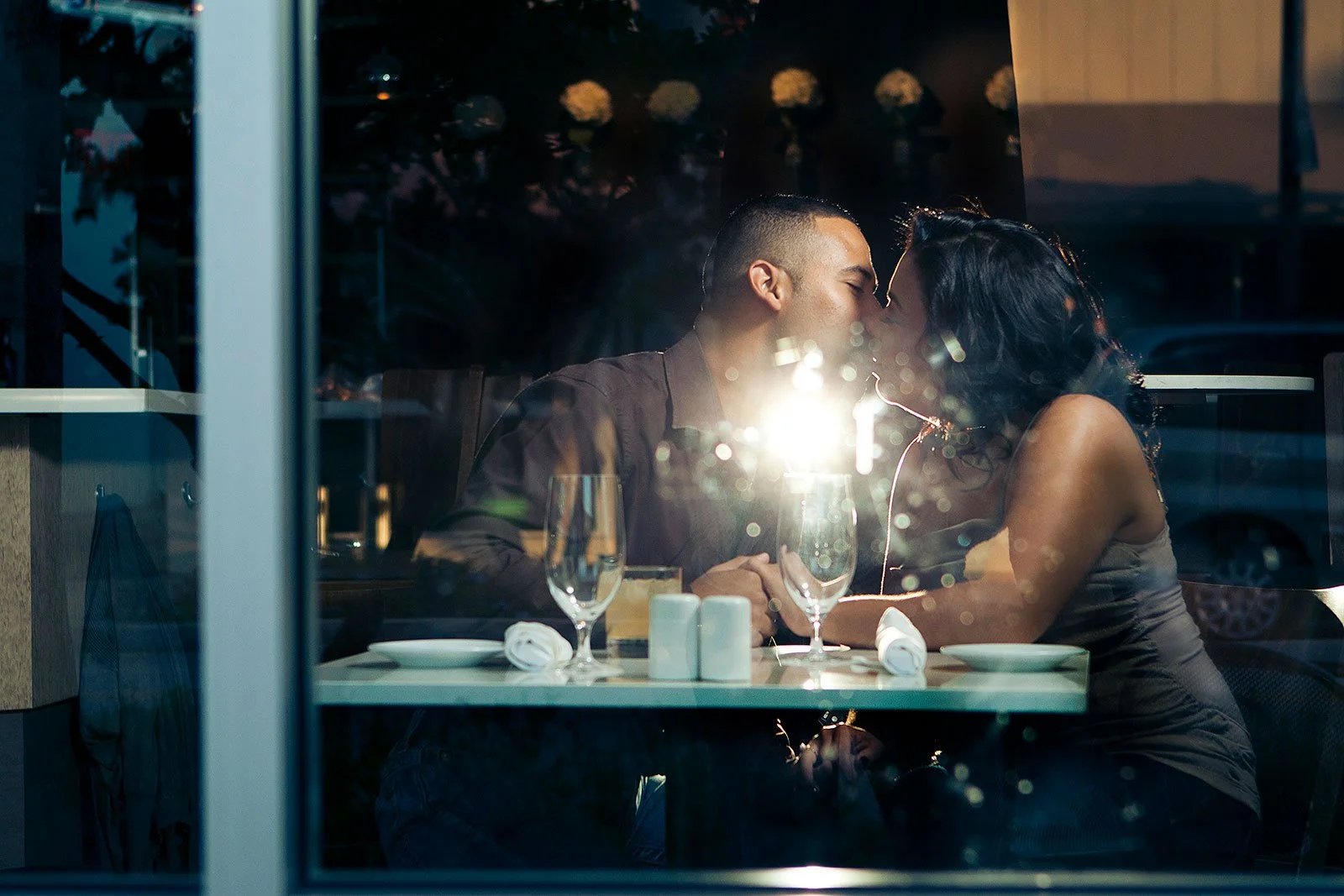 A couple sharing a kiss at a restaurant table, seen through a window with reflections, with empty wine glasses and table settings.